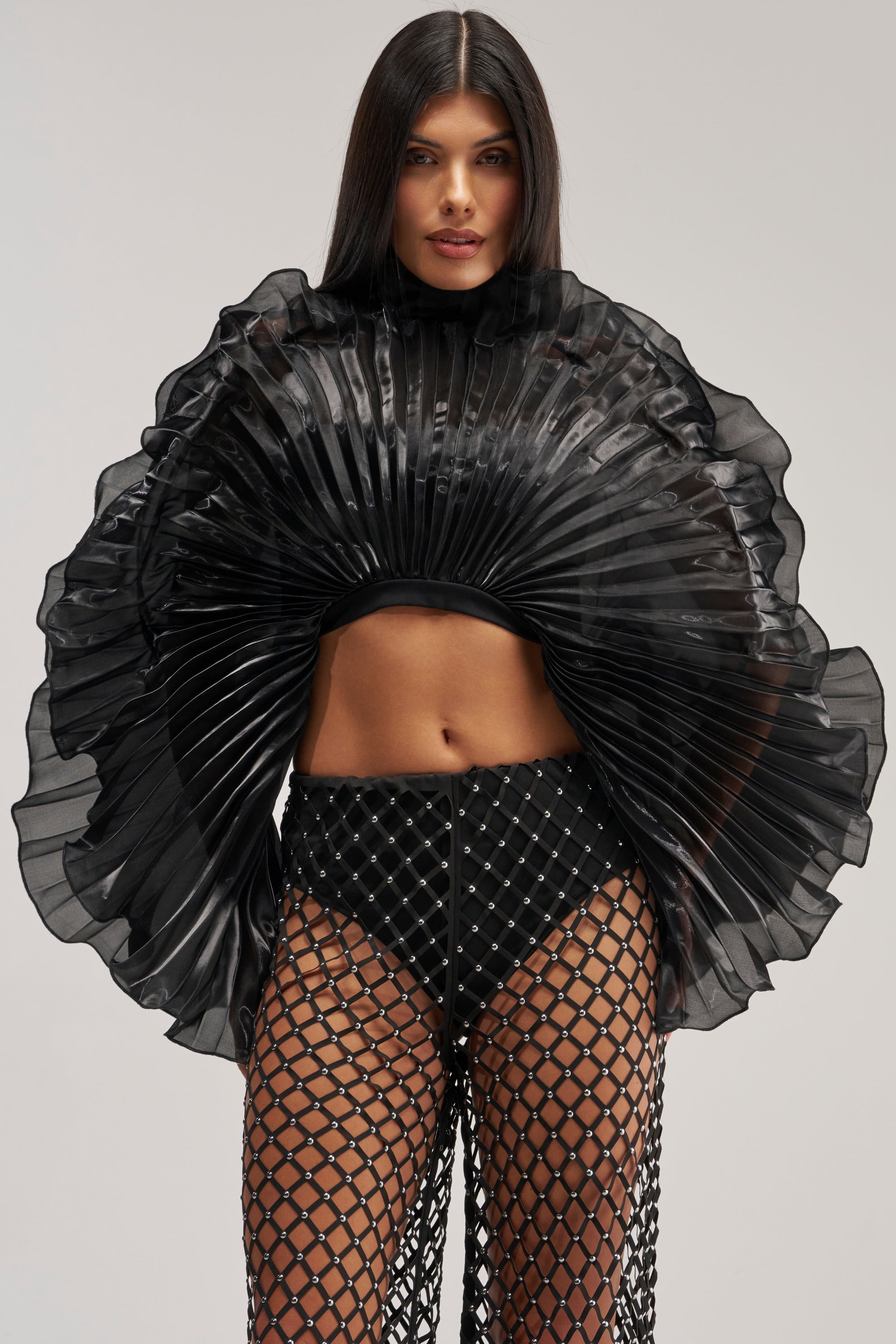 A woman models the SHINING DOWN ON YOU BLOUSE IN BLACK, a dramatic sheer pleated top featuring wide, fan-like sleeves, paired with matching black fishnet pants layered over high-waisted shorts. With her long, straight hair and neutral expression, she showcases this striking evening look in avant garde style.