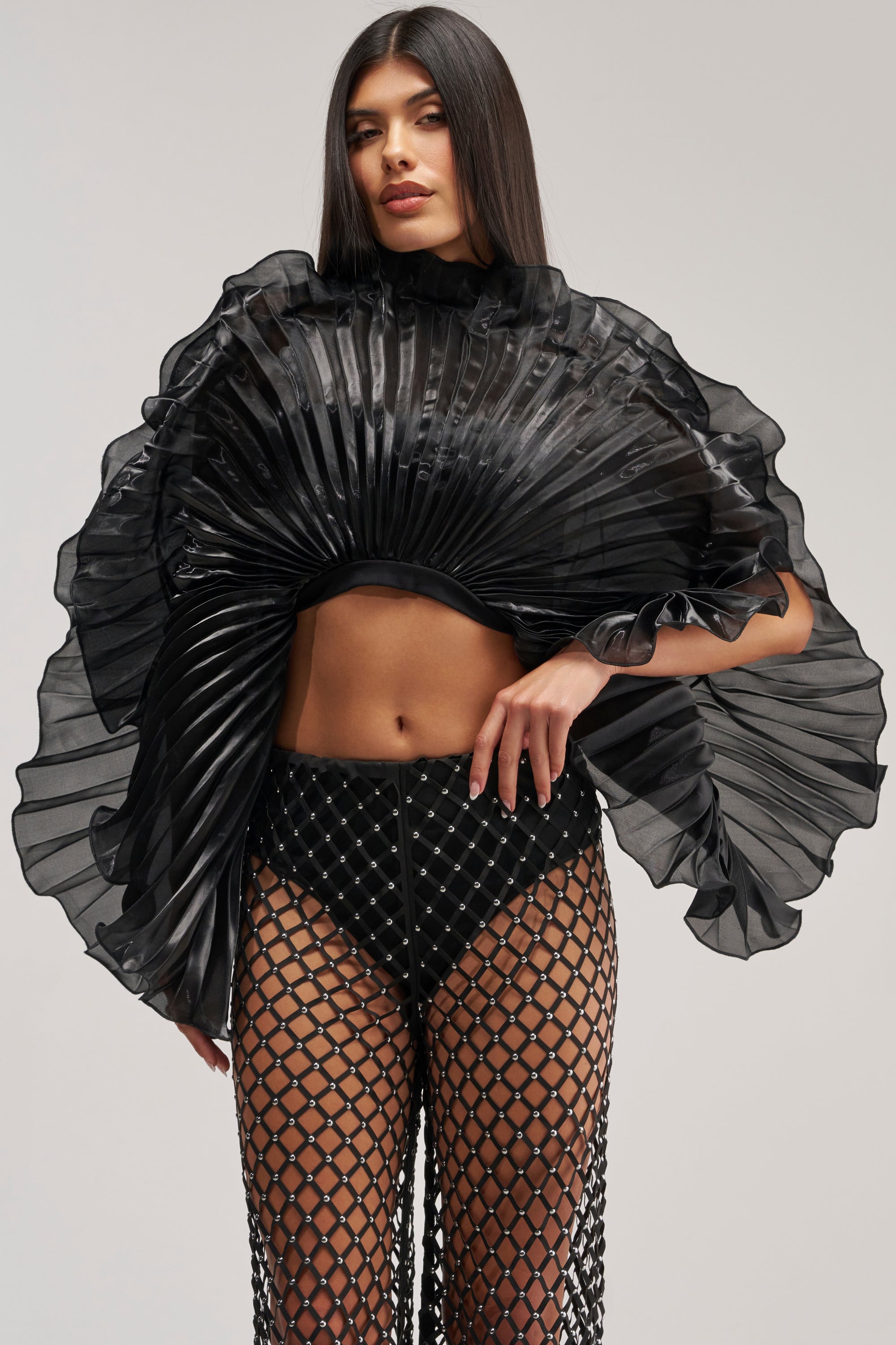 Wearing the SHINING DOWN ON YOU BLOUSE IN BLACK—a dramatic, avant garde sheer pleated top with ruffled sleeves—paired with high-waisted black mesh netted pants over black underwear, the woman stands against a plain background.
