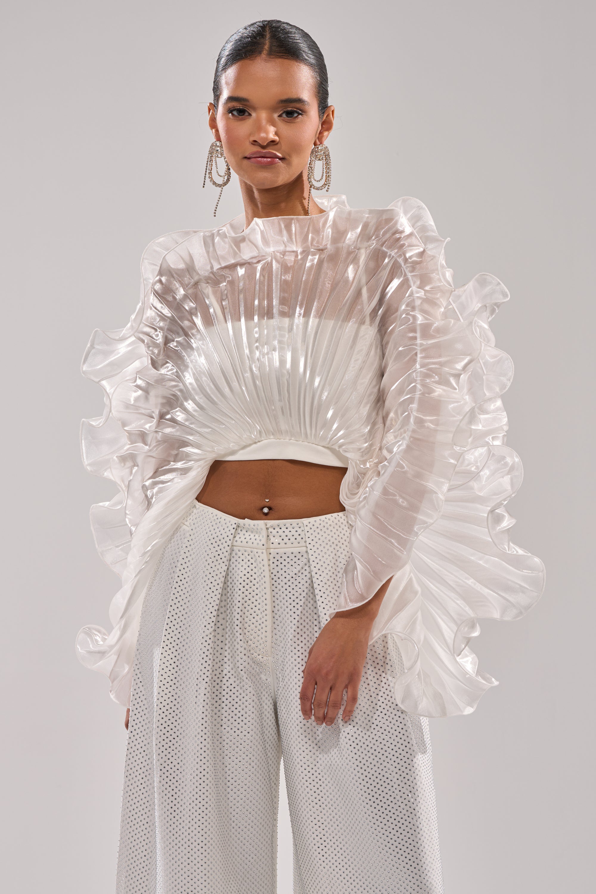 A woman wears the SHINING DOWN ON YOU BLOUSE with architectural ruffles and texture, paired with high-waisted white wide-leg pants. Her hair is slicked back, she wears statement earrings, and stands against a plain background. 0
