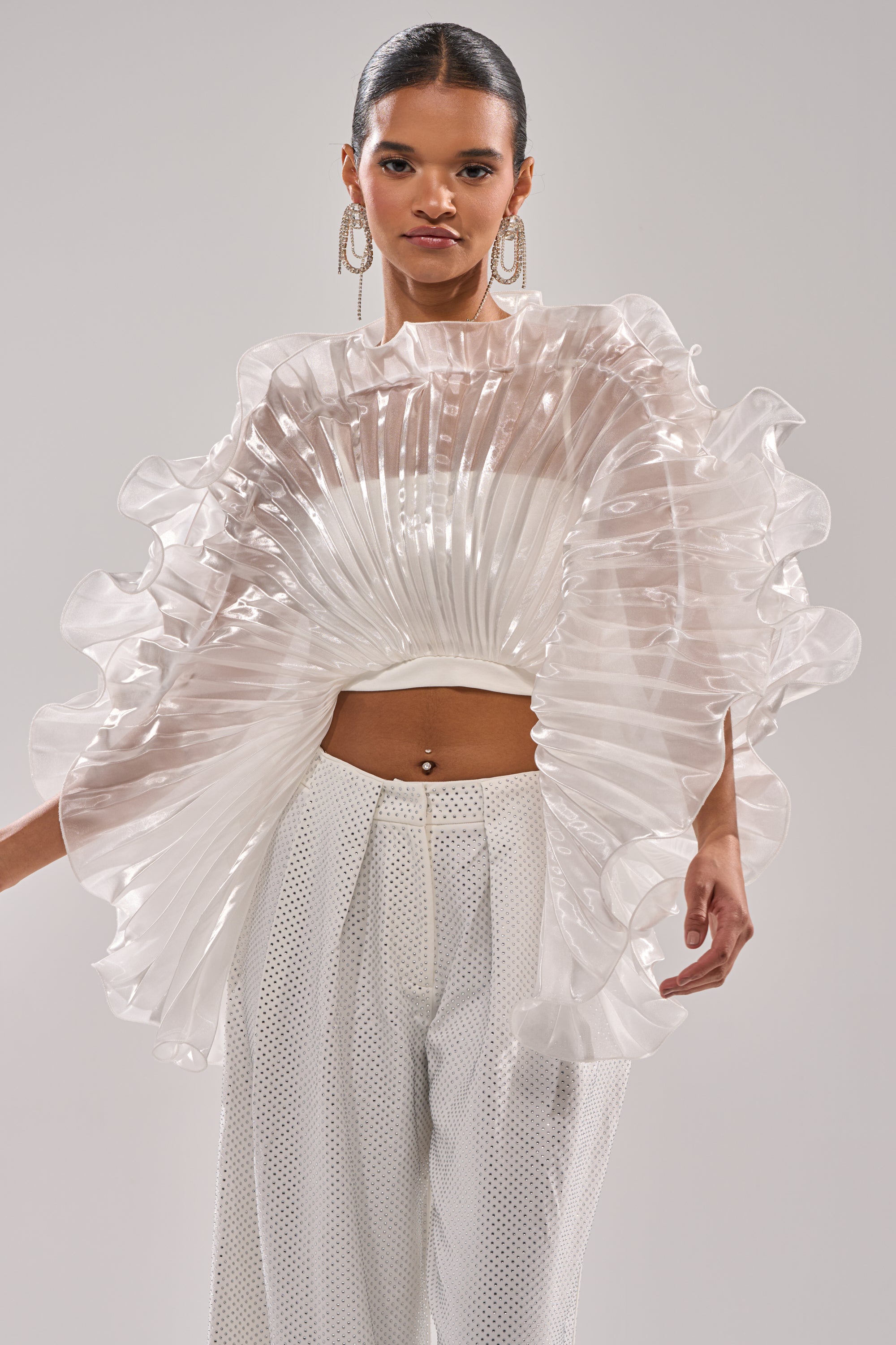 A woman poses in the SHINING DOWN ON YOU BLOUSE, featuring dramatic, sculptural ruffle sleeves, paired with white perforated pants. She accessorizes with large hoop earrings and slicked-back hair against a plain gray background. 2
