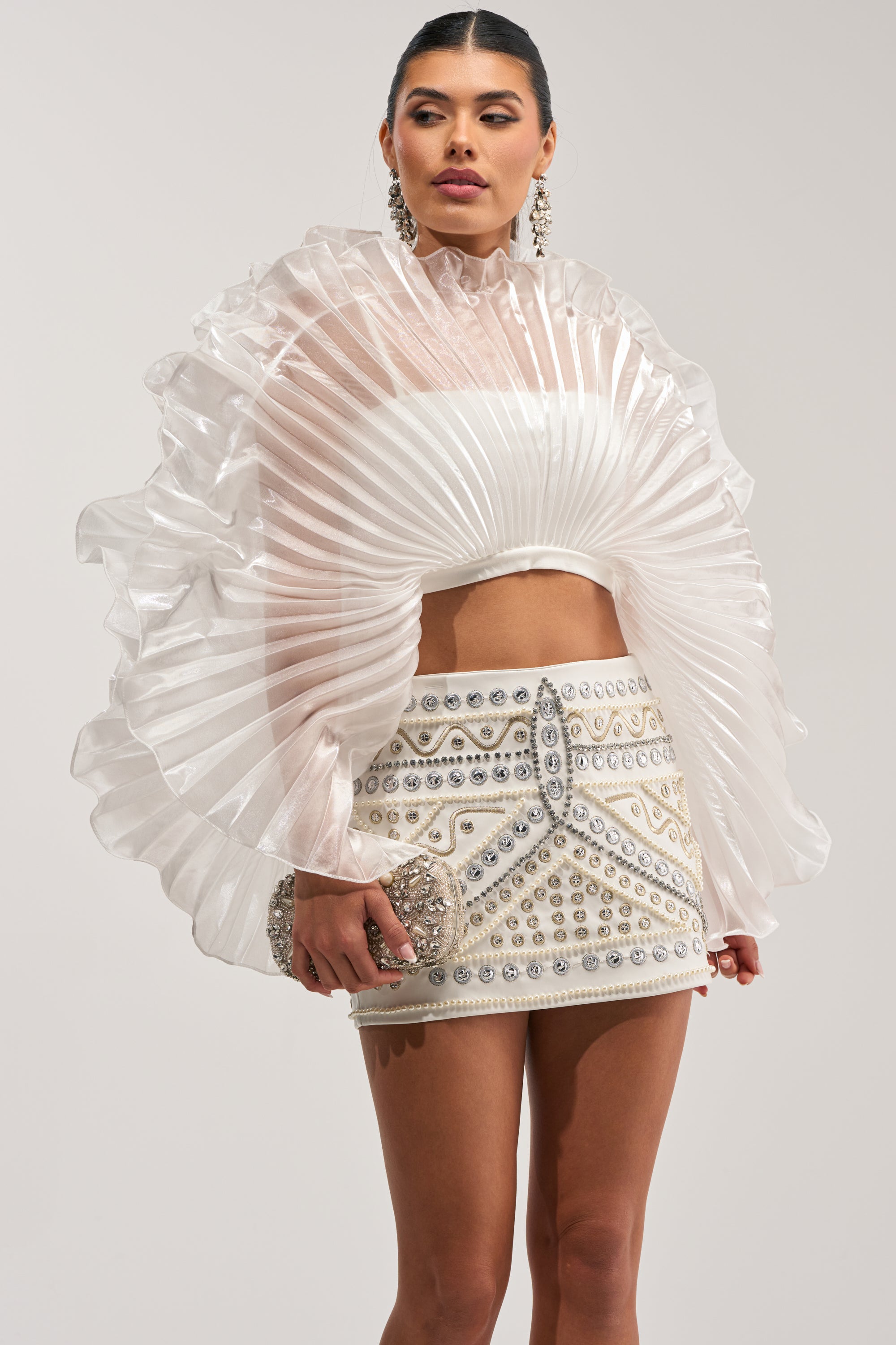 Wearing the SHINING DOWN ON YOU BLOUSE—a sheer, pleated white top with ruffled sleeves—paired with a short embellished skirt, she accessorizes with a sparkling clutch. Her hair is slicked back and she wears large, dangling earrings.