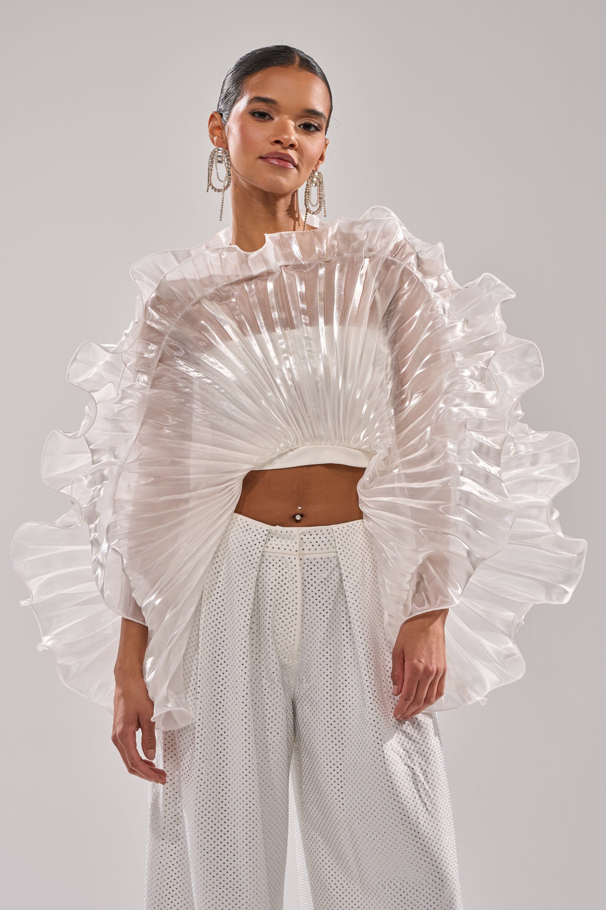 A woman wears the SHINING DOWN ON YOU BLOUSE with wide sleeves and white high-waisted pants, paired with statement earrings. She strikes a confident pose against a minimalist background. 3