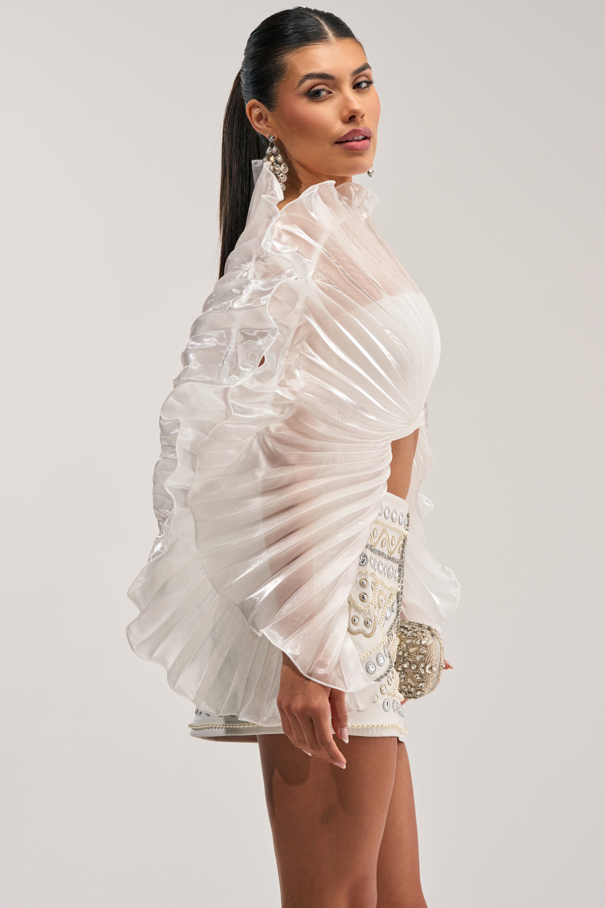 A woman with a high ponytail and glamorous makeup poses in profile, wearing the SHINING DOWN ON YOU BLOUSE—an avant-garde evening top defined by its voluminous, pleated ruffled sleeves—paired with a matching miniskirt intricately embellished with pearls and crystals, and holding a coordinating round clutch.