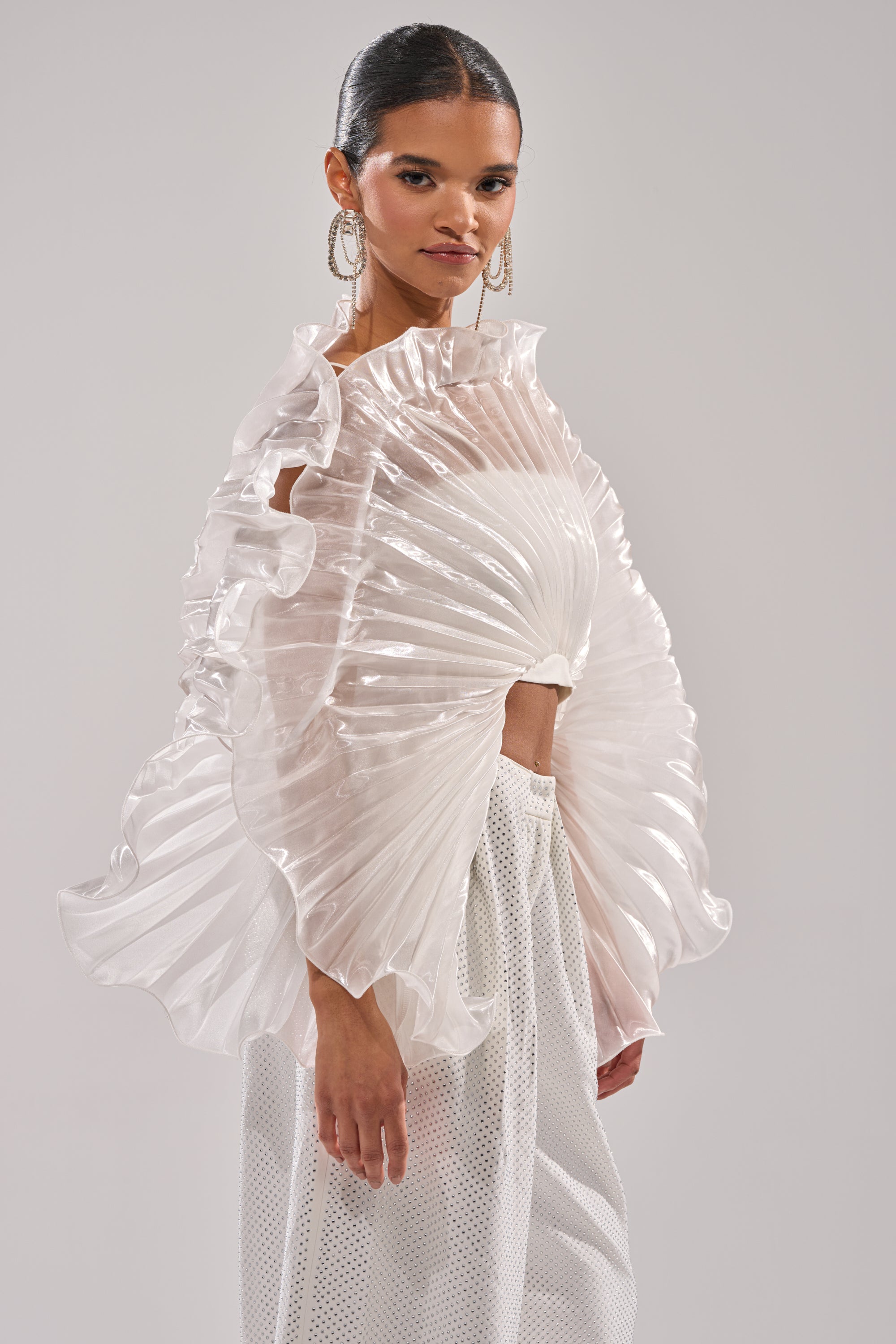 A woman models the SHINING DOWN ON YOU BLOUSE, featuring sheer sculptural pleats, dramatic ruffled edges, and wide sleeves, styled with white textured pants. Her hair is sleek and tied back, with large hoop earrings completing the look. 4