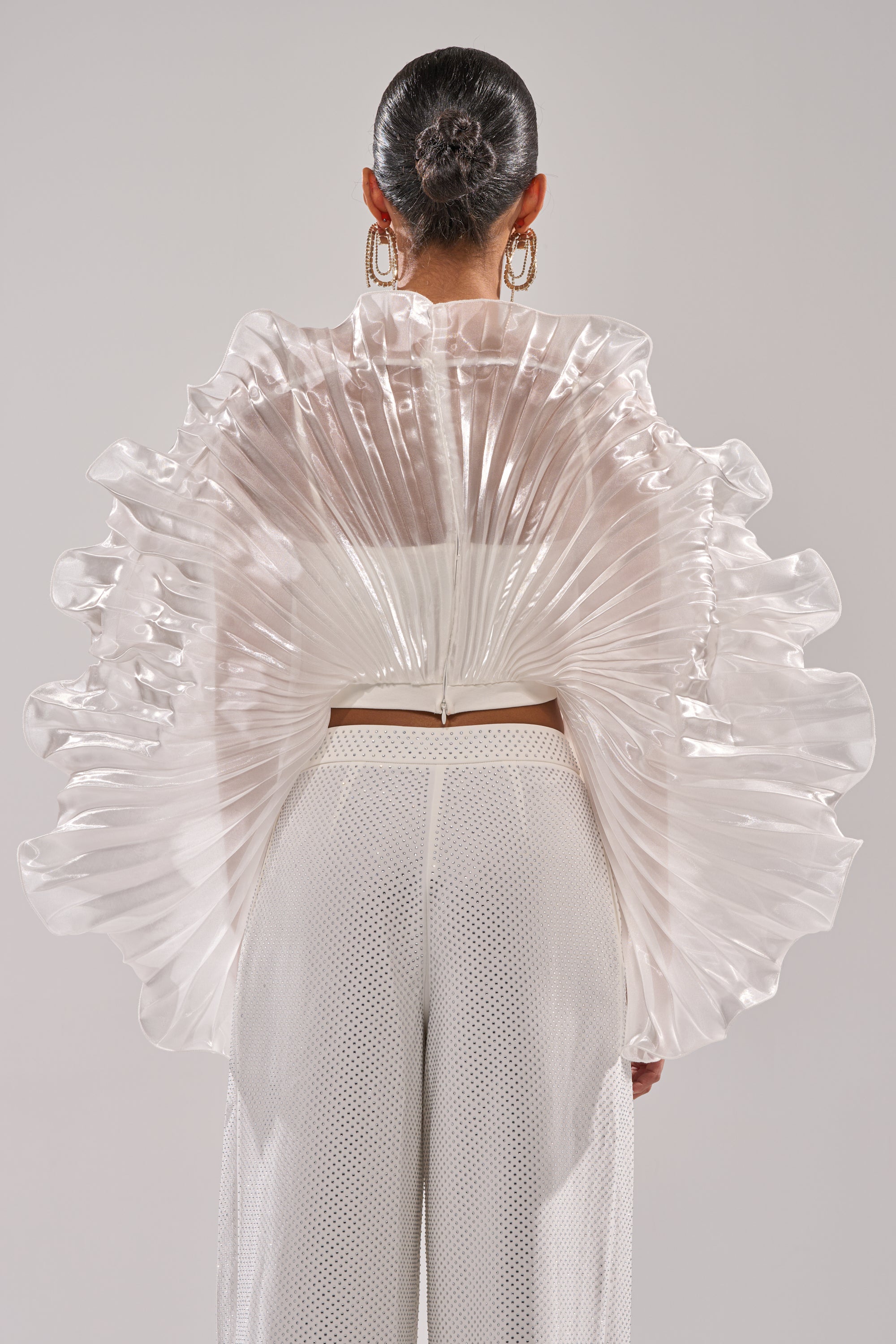 A person seen from behind wears the SHINING DOWN ON YOU BLOUSE, featuring sculptural, translucent pleats that fan out dramatically. They pair it with high-waisted white pants and accessorize with a sleek bun and large hoop earrings. 5