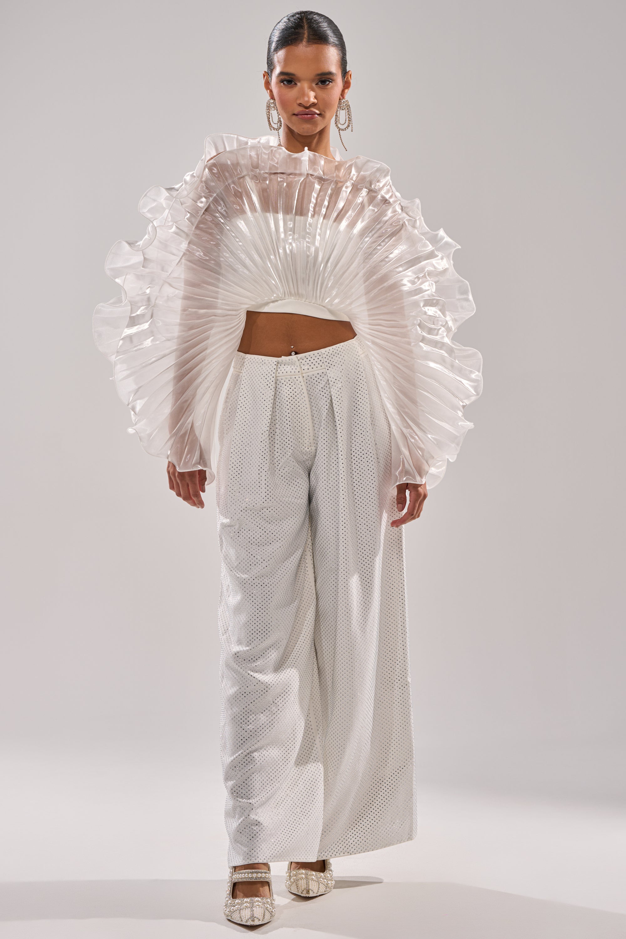 A woman stands facing forward in the SHINING DOWN ON YOU BLOUSE, featuring architectural ruffles and long sleeves, paired with high-waisted, wide-leg white pants. Her hair is pulled back and she accessorizes with hoop earrings and pointed shoes against a plain background. 6
