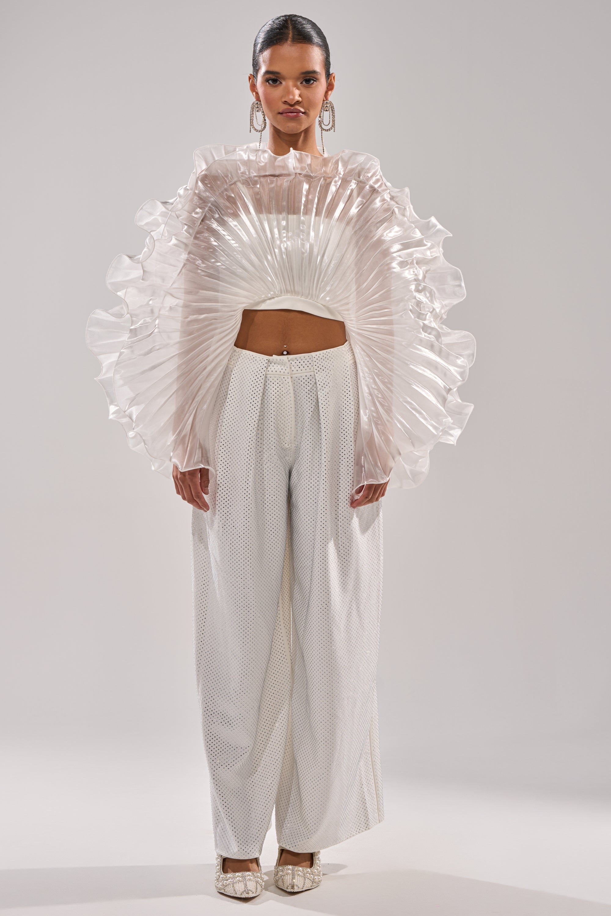 A woman stands facing forward in wide-legged white pants and the SHINING DOWN ON YOU BLOUSE, featuring voluminous sleeves and architectural ruffles. She pairs hoop earrings, pulled-back hair, and neutral heels against a plain light background. 7