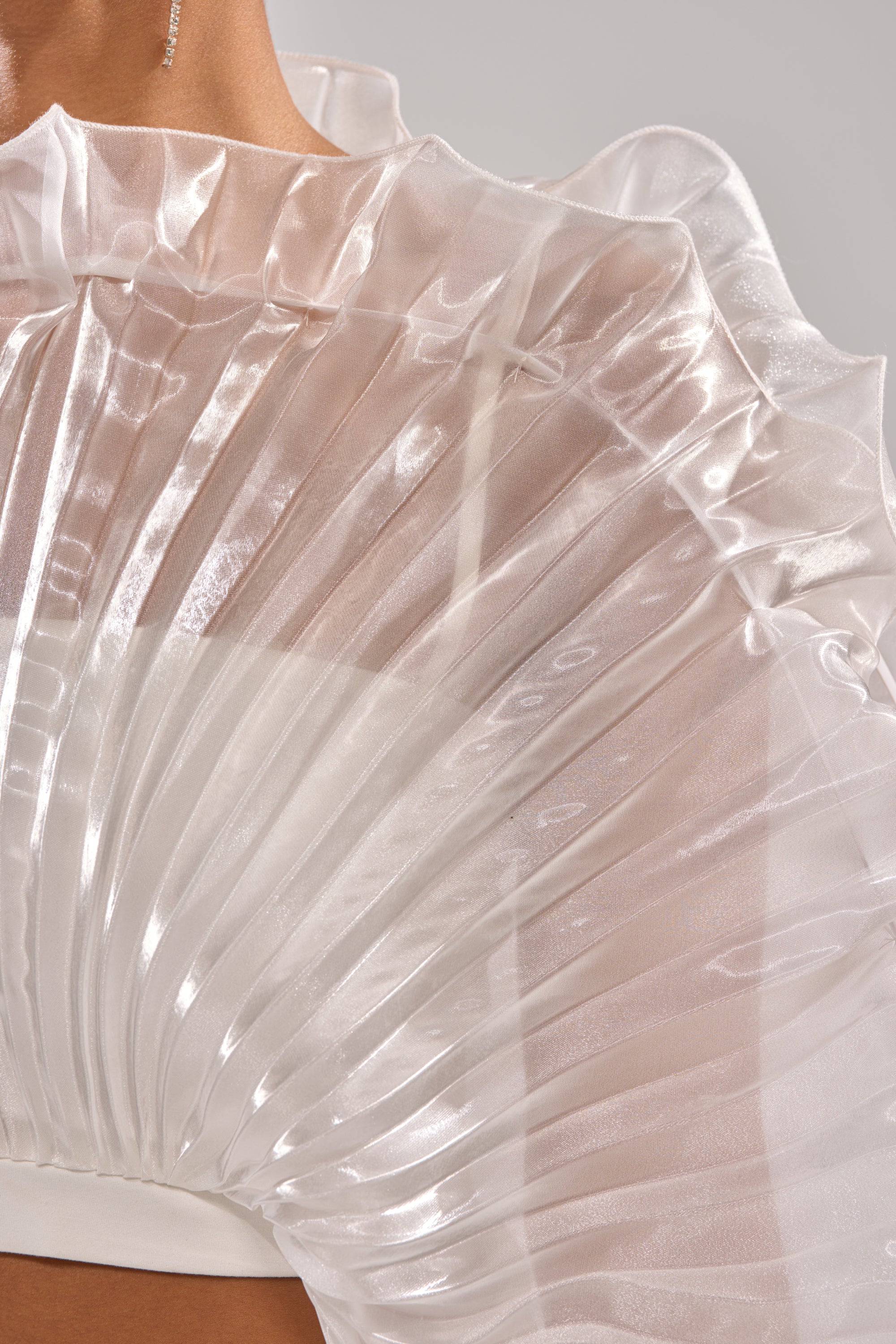 Close-up of a person wearing the SHINING DOWN ON YOU BLOUSE, featuring sheer, pleated iridescent white fabric and ruffled edges draped over the shoulder for a dramatic, elegant look. Part of the neck and shoulder is visible. 8