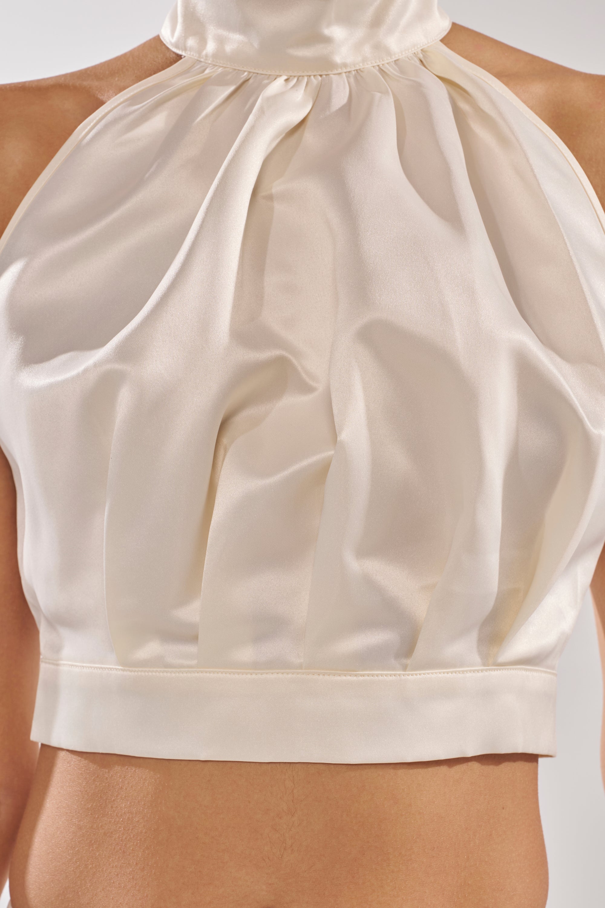 A close-up of a person wearing the IT WAS EASY MOCKNECK BLOUSE—a sleeveless, ivory satin crop top with pleated front detailing—perfect as an elegant bridal choice. 9
