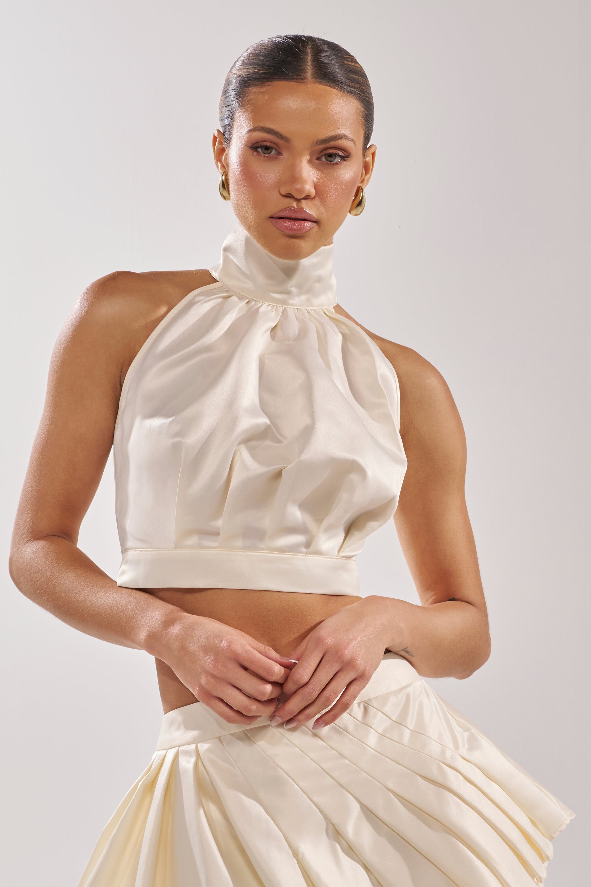 A woman poses against a plain light background wearing the IT WAS EASY MOCKNECK BLOUSE—an ivory crop top with a gathered front and high neck—paired with a matching pleated cream skirt. 3