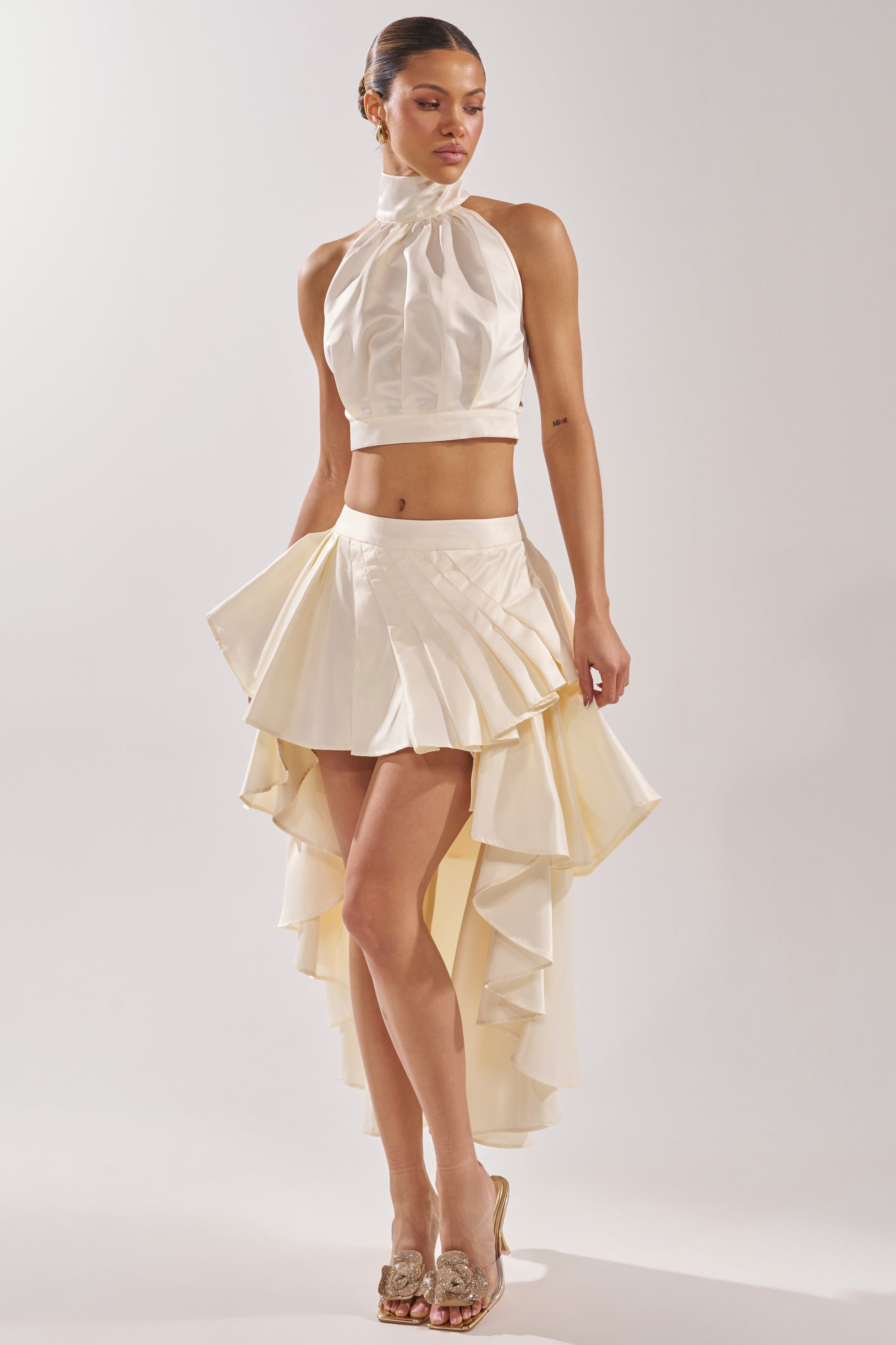 She wears the IT WAS EASY MOCKNECK BLOUSE in ivory with an asymmetrical cream skirt, featuring a short front and long, ruffled back. Paired with heels, she stands elegantly against a plain background. 6