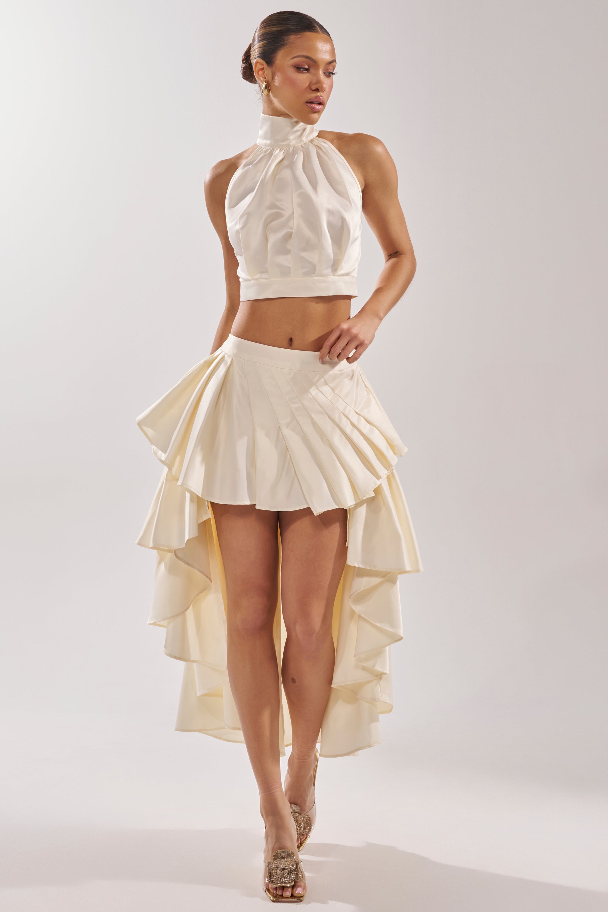 A woman models the IT WAS EASY MOCKNECK BLOUSE in ivory, paired with a matching high-low ruffle skirt and open-toe heels. She stands against a plain white background, showcasing a chic bridal crop top look. 7