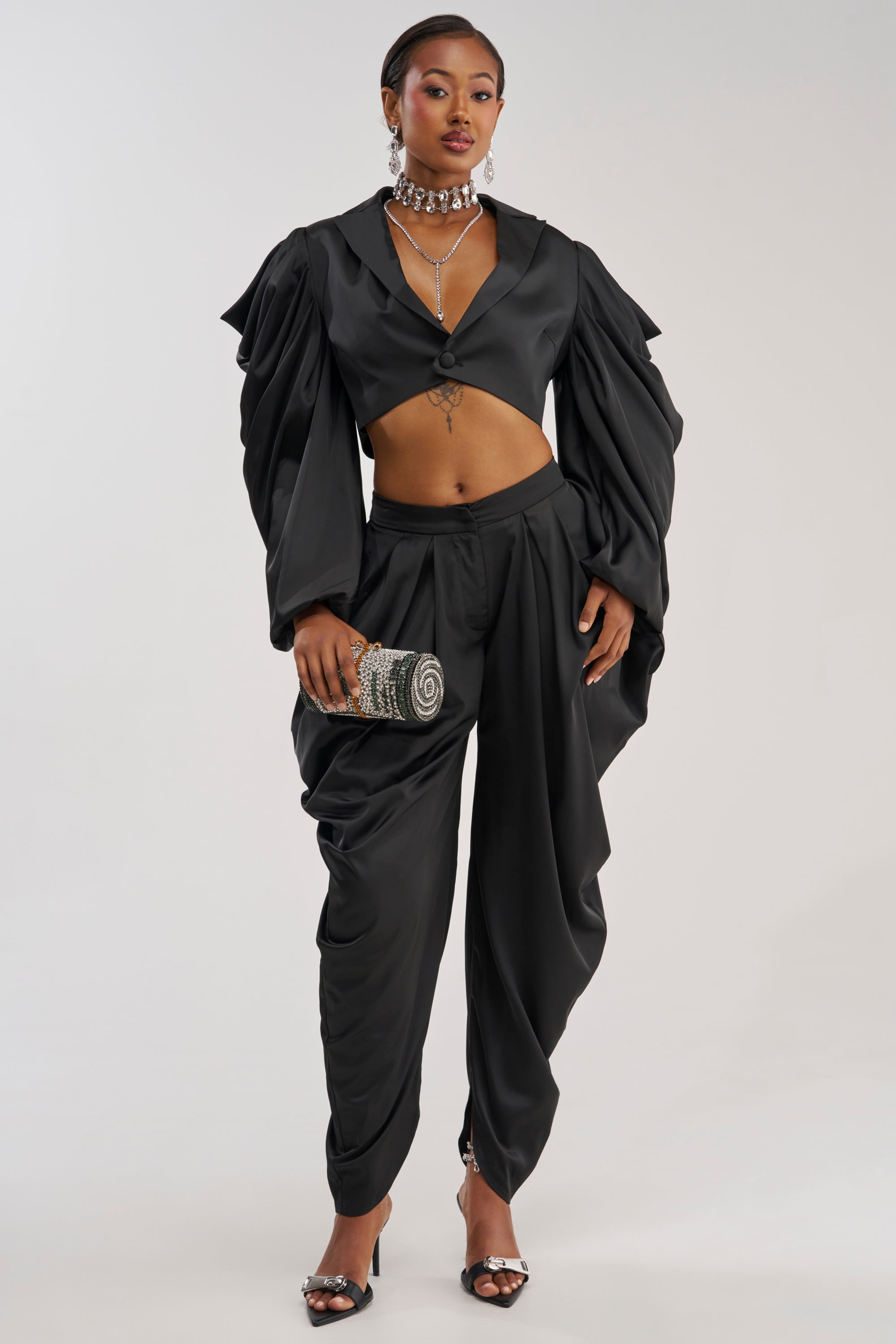 A person wearing the DRIP DRIP TROUSER IN BLACK, paired with a black satin crop top featuring dramatic sleeves, accessorized with a jeweled clutch, choker necklace, earrings, and open-toed heels, stands against a plain light background.