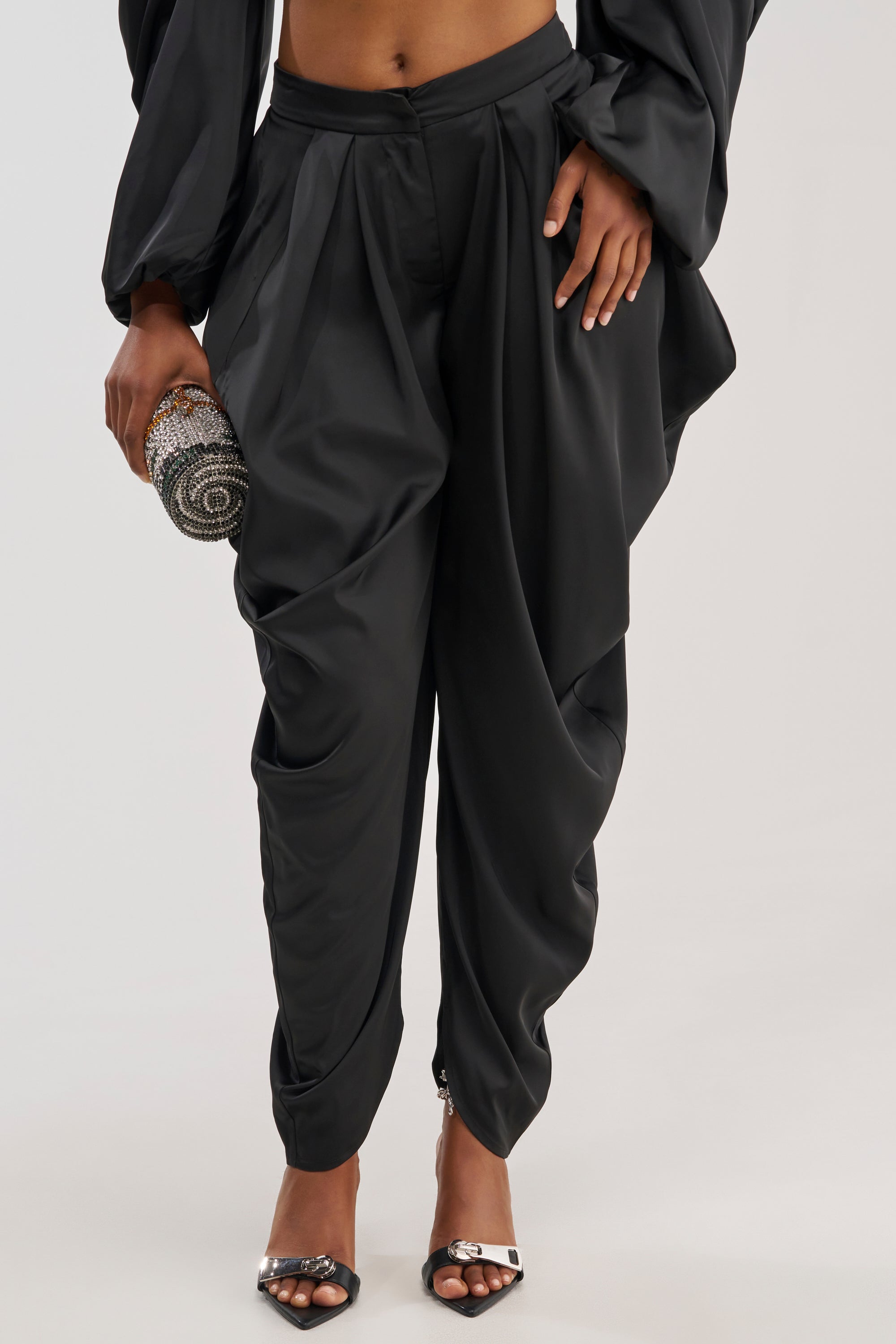 A person wearing the DRIP DRIP TROUSER IN BLACK, paired with black heels featuring metallic accents and holding a sparkly, round clutch. The photo is cropped from the shoulders down against a light background.