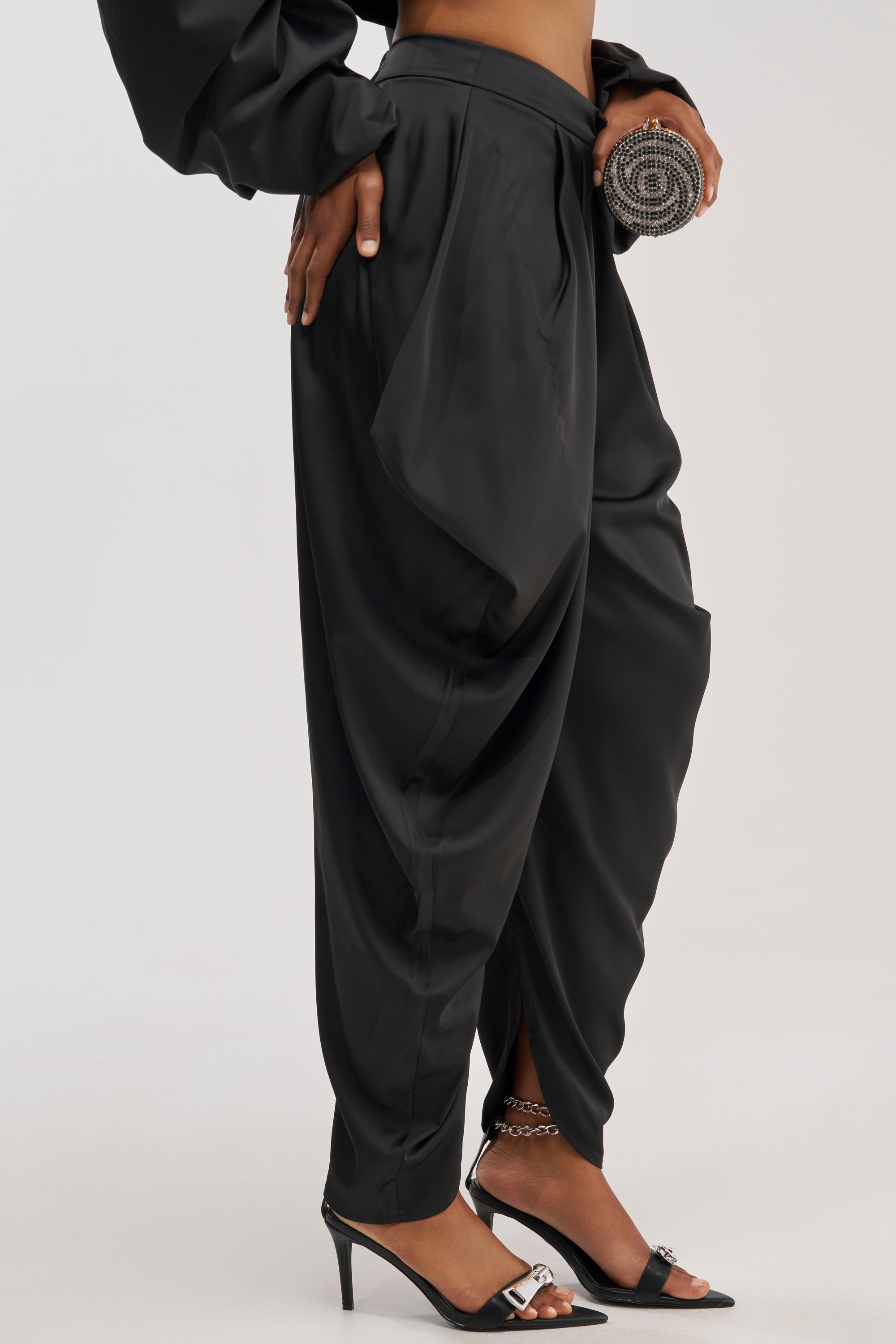 A person wearing the DRIP DRIP TROUSER IN BLACK, which features a wide-leg silhouette, paired with black ankle-chain heels and holding a small, round beaded clutch. Only the lower body and part of one arm are visible.