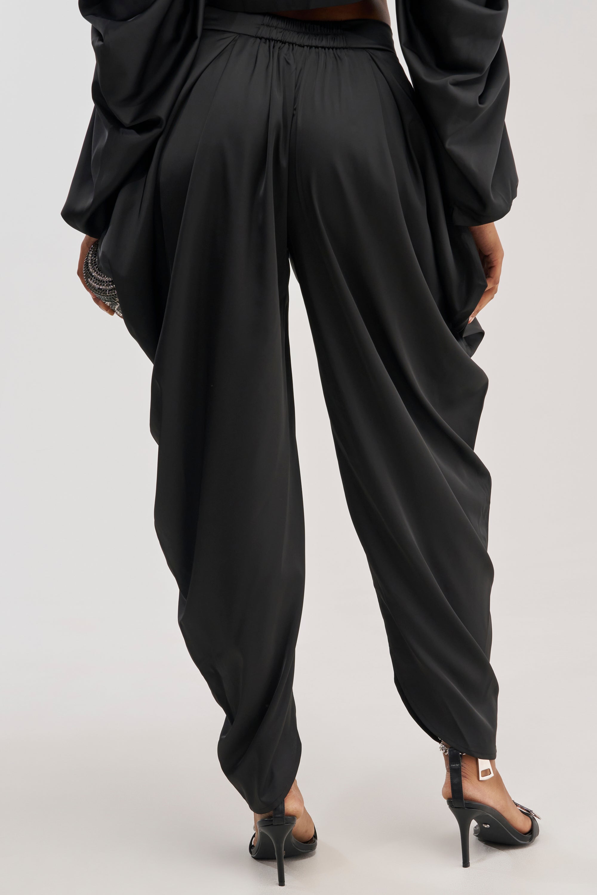 A person is seen from the back wearing the DRIP DRIP TROUSER IN BLACK—loose, draped black satin trousers with a wide-leg silhouette. Their outfit features long sleeves, complemented by black high-heeled sandals, and the flowing fabric lends an elegant, formal touch.