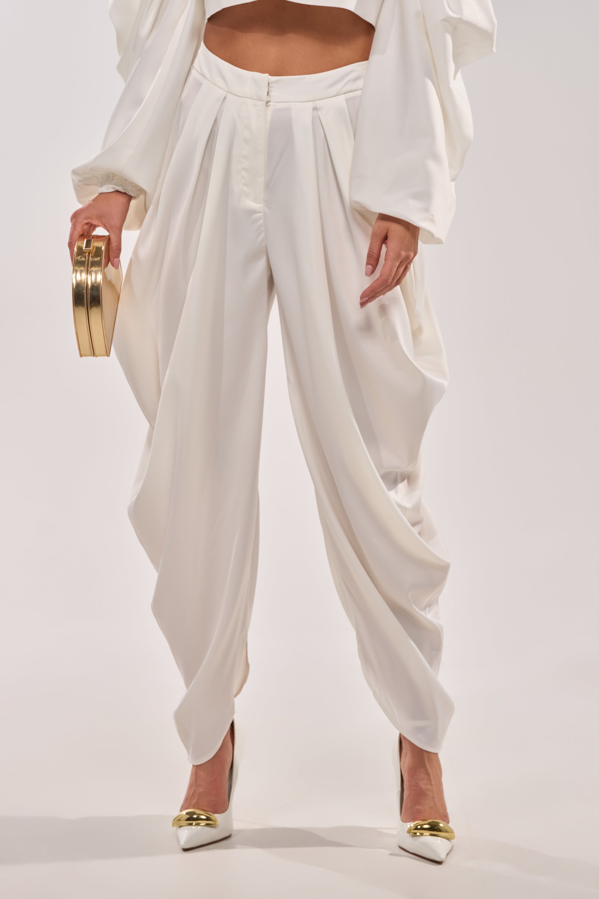 Wearing the NEW BEGINNING TROUSER with a matching white top, gold-tipped white heels, and a small gold clutch, a person stands against a plain light background. 1