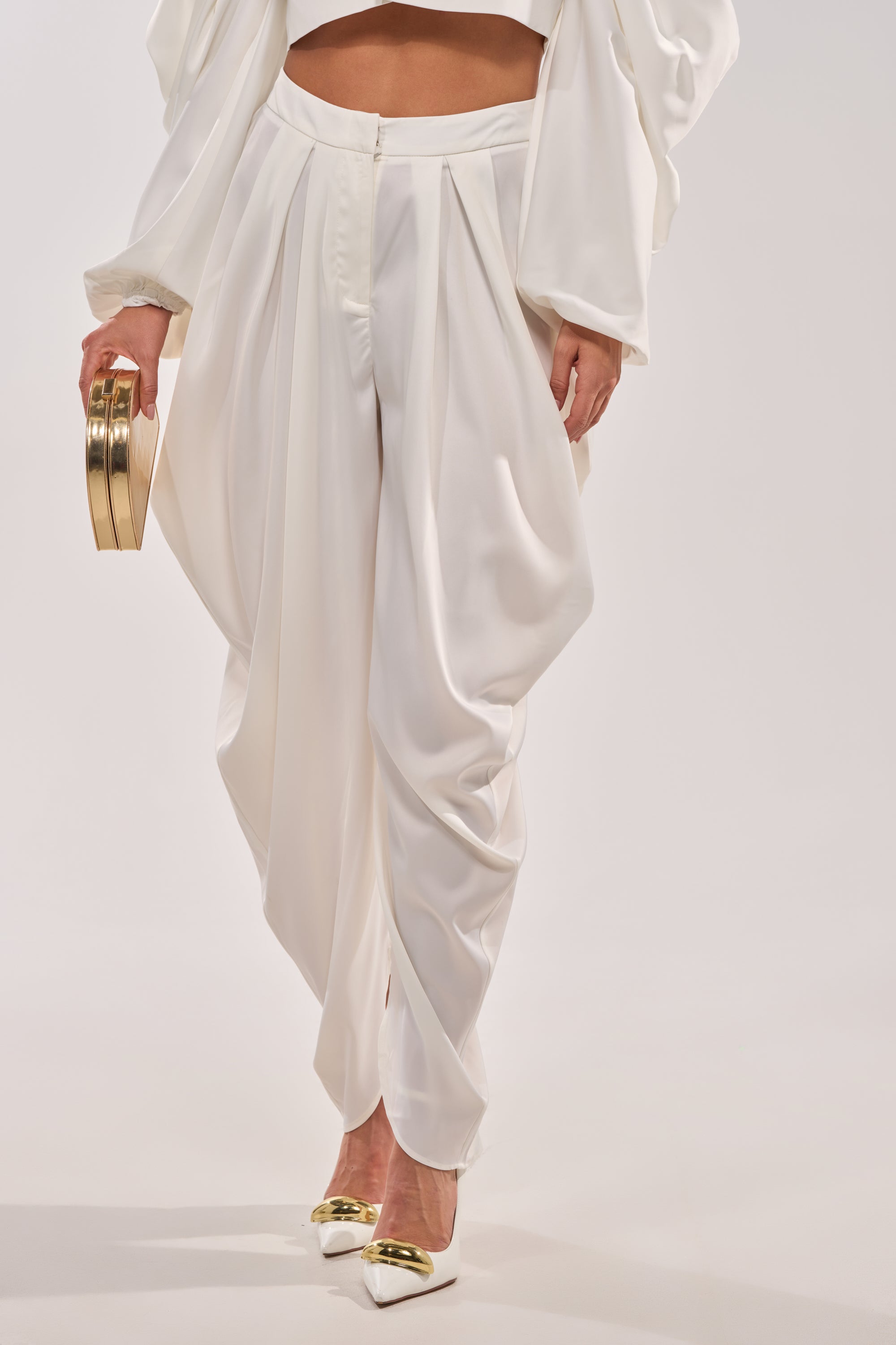 A person wears the NEW BEGINNING TROUSER in elegant white with a matching top, white heels with gold tips, and holds a round gold clutch, standing against a plain white background. 2
