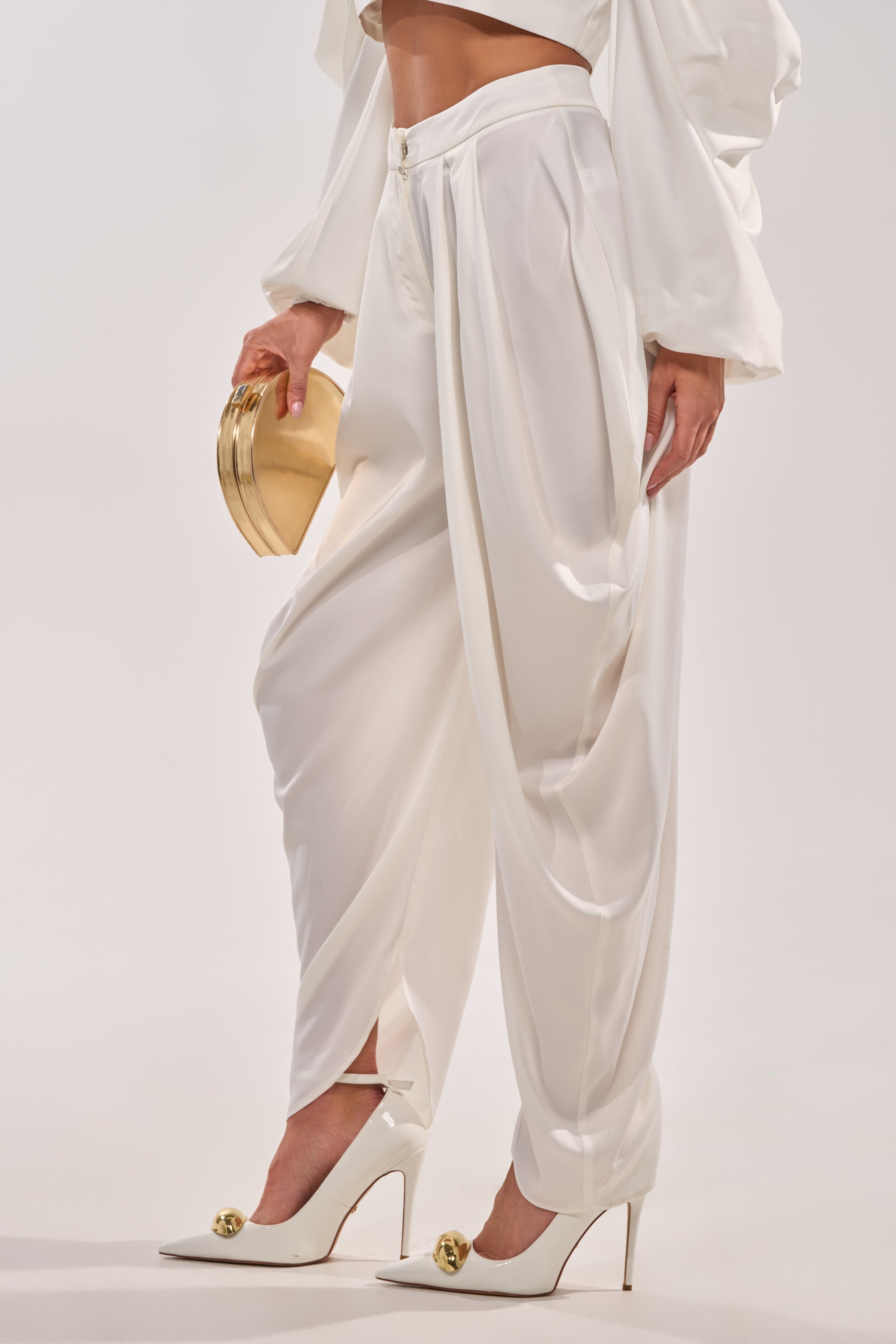A person wears the NEW BEGINNING TROUSER—elegant high-waisted draped pants—paired with white high heels with gold accents and a small gold clutch, set against a plain light background. 3