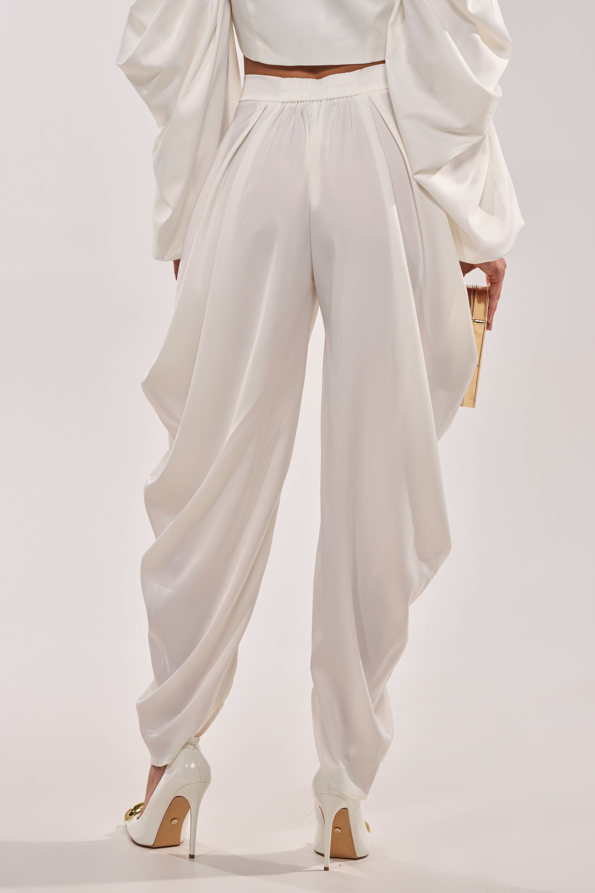 Wearing the NEW BEGINNING TROUSER in ivory with a matching long-sleeve top and white heels, a person holds a small gold clutch. The photo is taken from behind against a plain background. 4