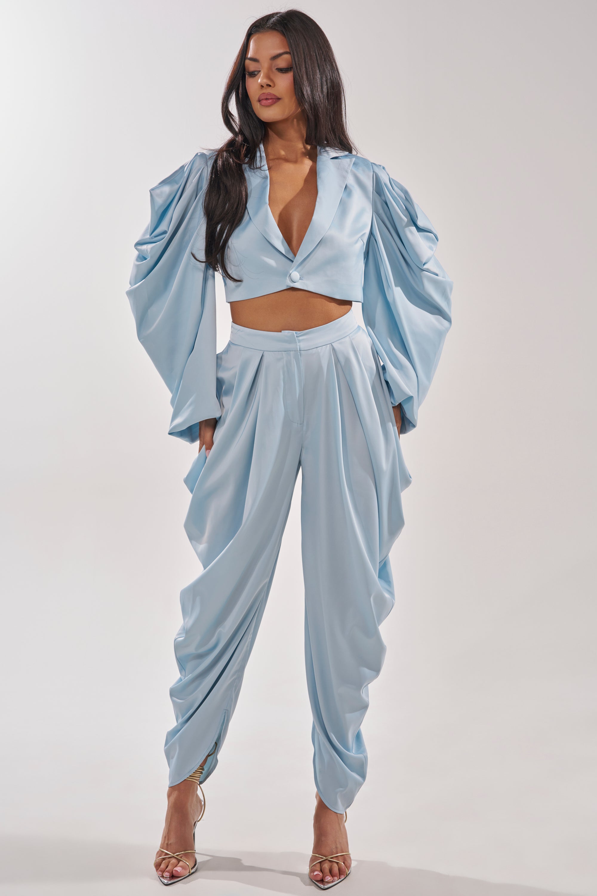 A woman wears the DRIP DRIP TROUSER, a shiny light blue high-rise satin pant paired with a dramatic draped sleeve top, standing in heels against a plain background.