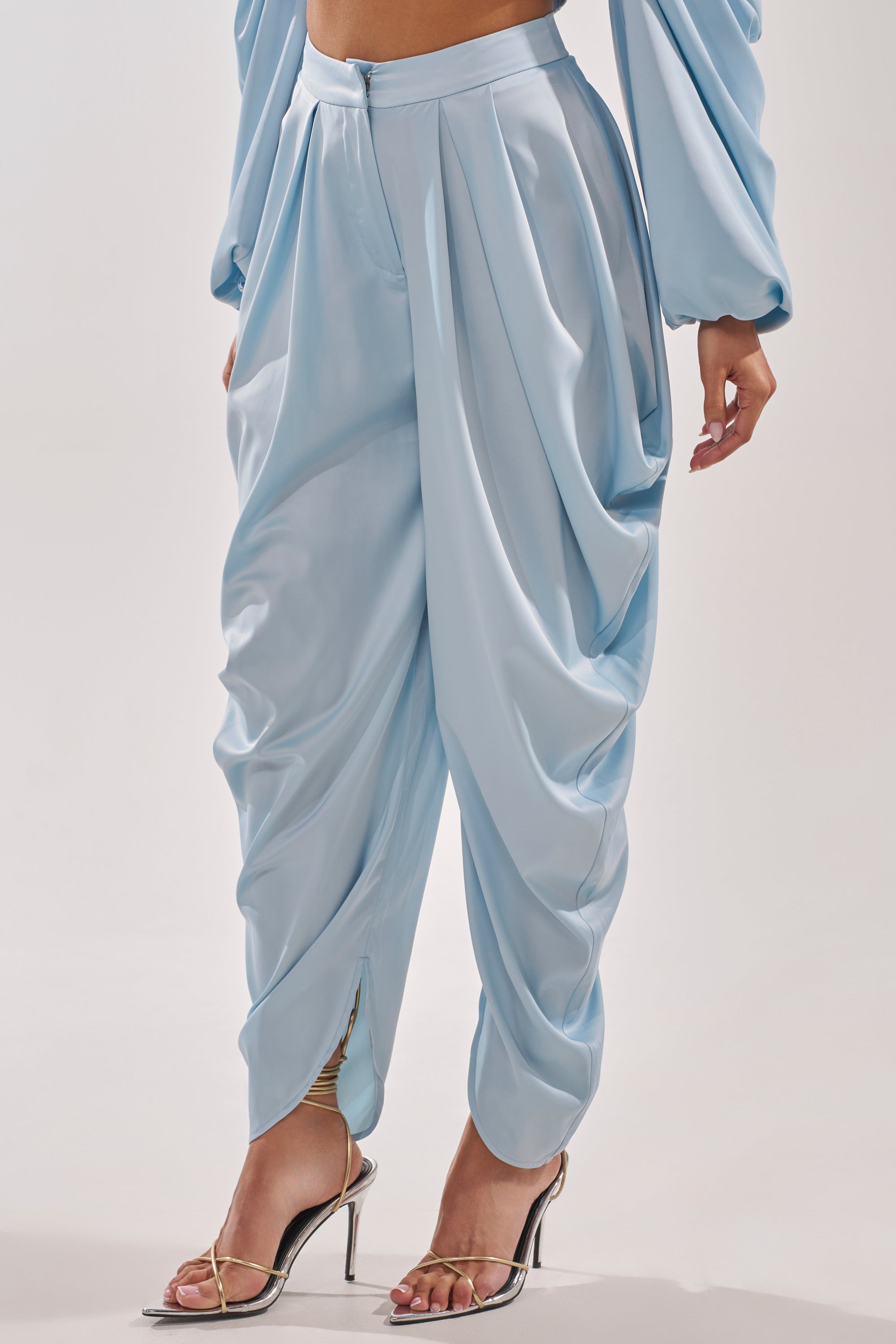 A person wears the DRIP DRIP TROUSER, featuring powder blue satin, a high-rise waist, and a loose, pleated harem-style design, paired with metallic silver high-heeled sandals against a plain light background.