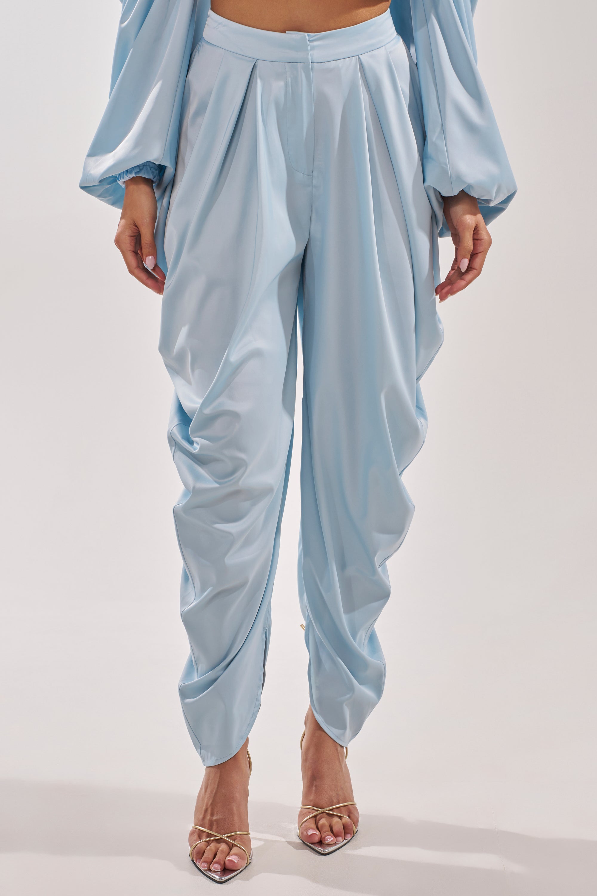 A person wears the DRIP DRIP TROUSER in powder blue satin with a high-rise waist, paired with a matching long-sleeve top and open-toe heeled sandals. Only the lower body is shown against a plain background.