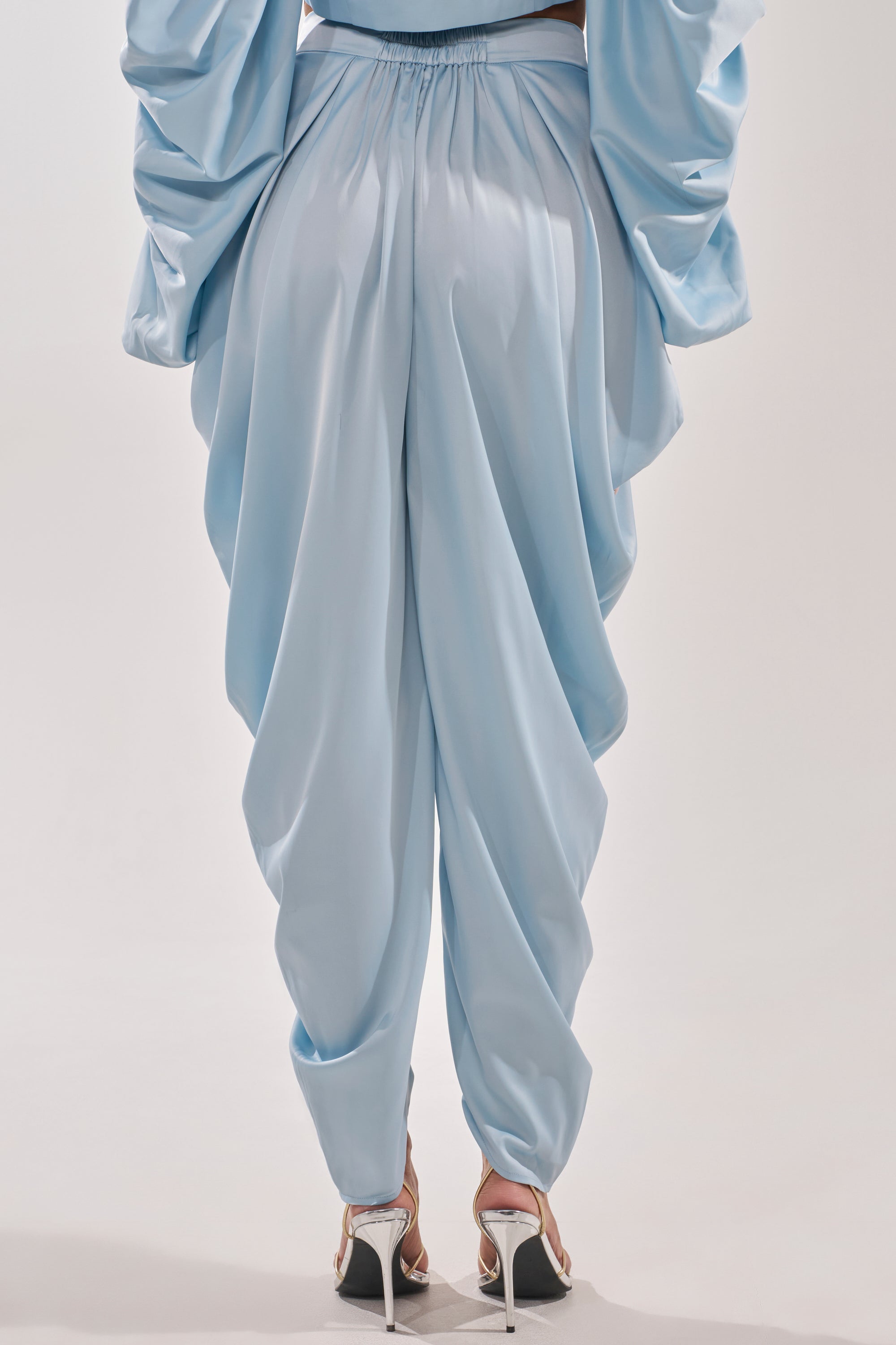 Someone wearing the DRIP DRIP TROUSER in powder blue satin, featuring a high-rise waist, paired with high-heeled sandals, shown from the back against a plain background.