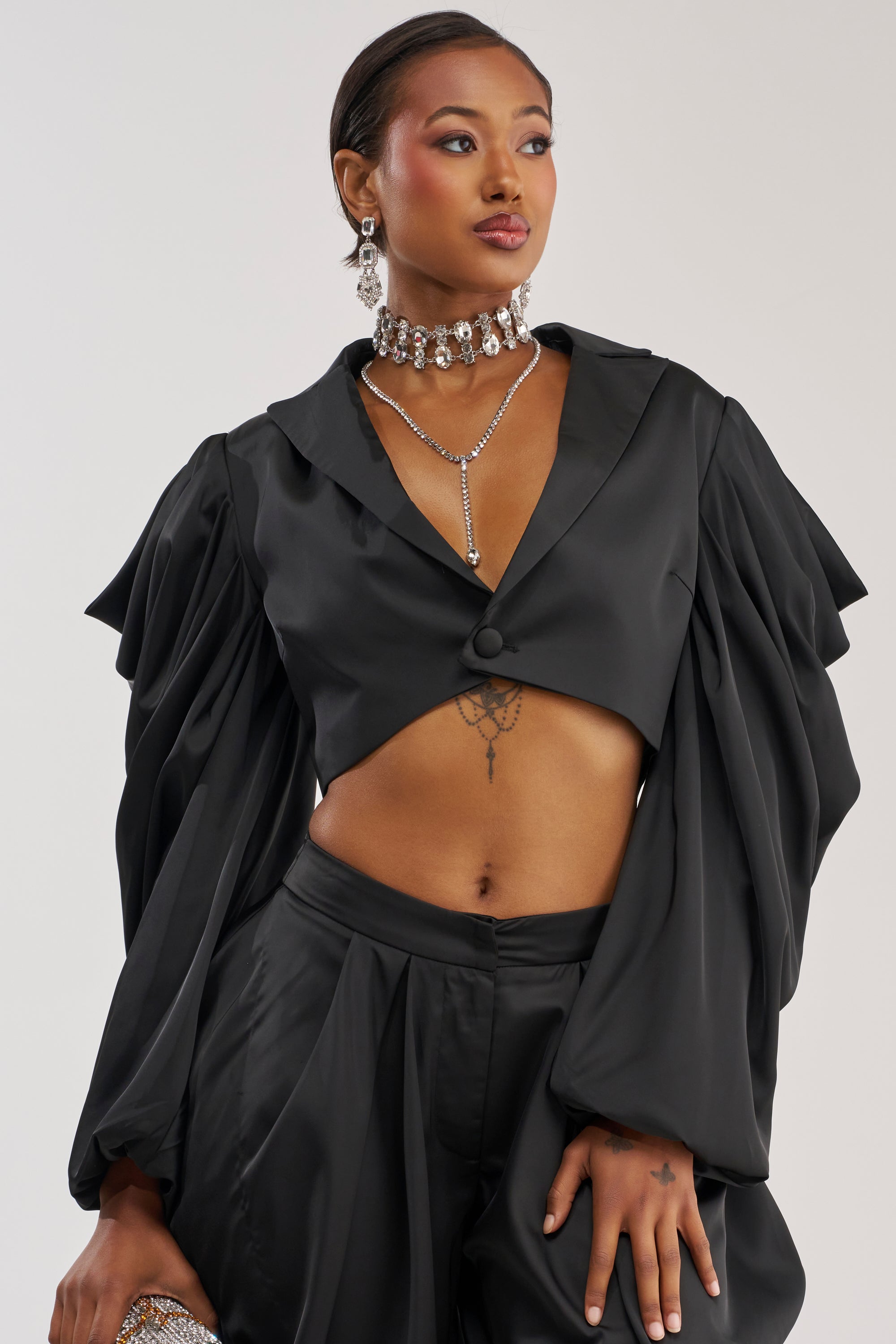 A woman wears the DRIP DRIP BLAZER IN BLACK, a satin cropped blazer featuring dramatic, draped sleeves, paired with matching pants. She accessorizes with a necklace and earrings, revealing a midriff tattoo as she gazes to the side against a plain background.