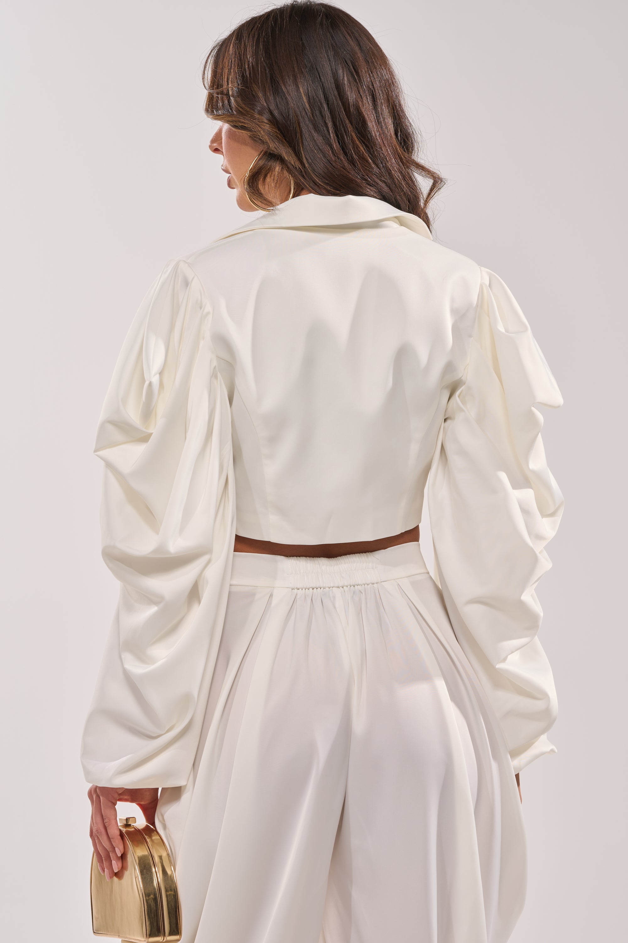 A woman stands with her back to the camera in the NEW BEGINNING CROPPED BLAZER, styled with ruched sleeves and matching high-waisted pants, holding a gold purse against a plain background. 4