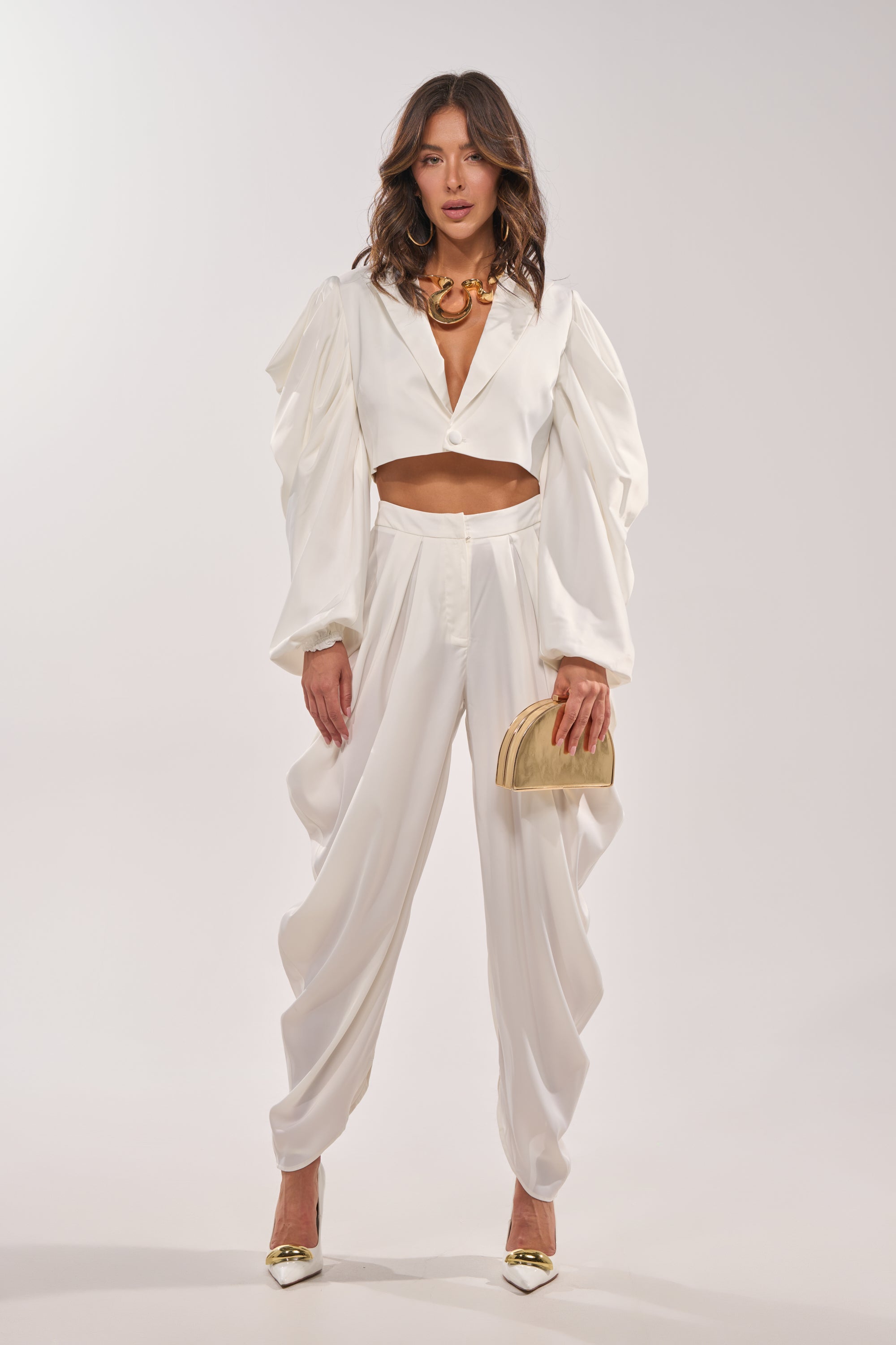 A woman stands facing forward, wearing the NEW BEGINNING CROPPED BLAZER with voluminous cape sleeves and high-waisted, draped white pants. She accessorizes with a beige clutch, gold heels, and a chunky gold necklace. 5