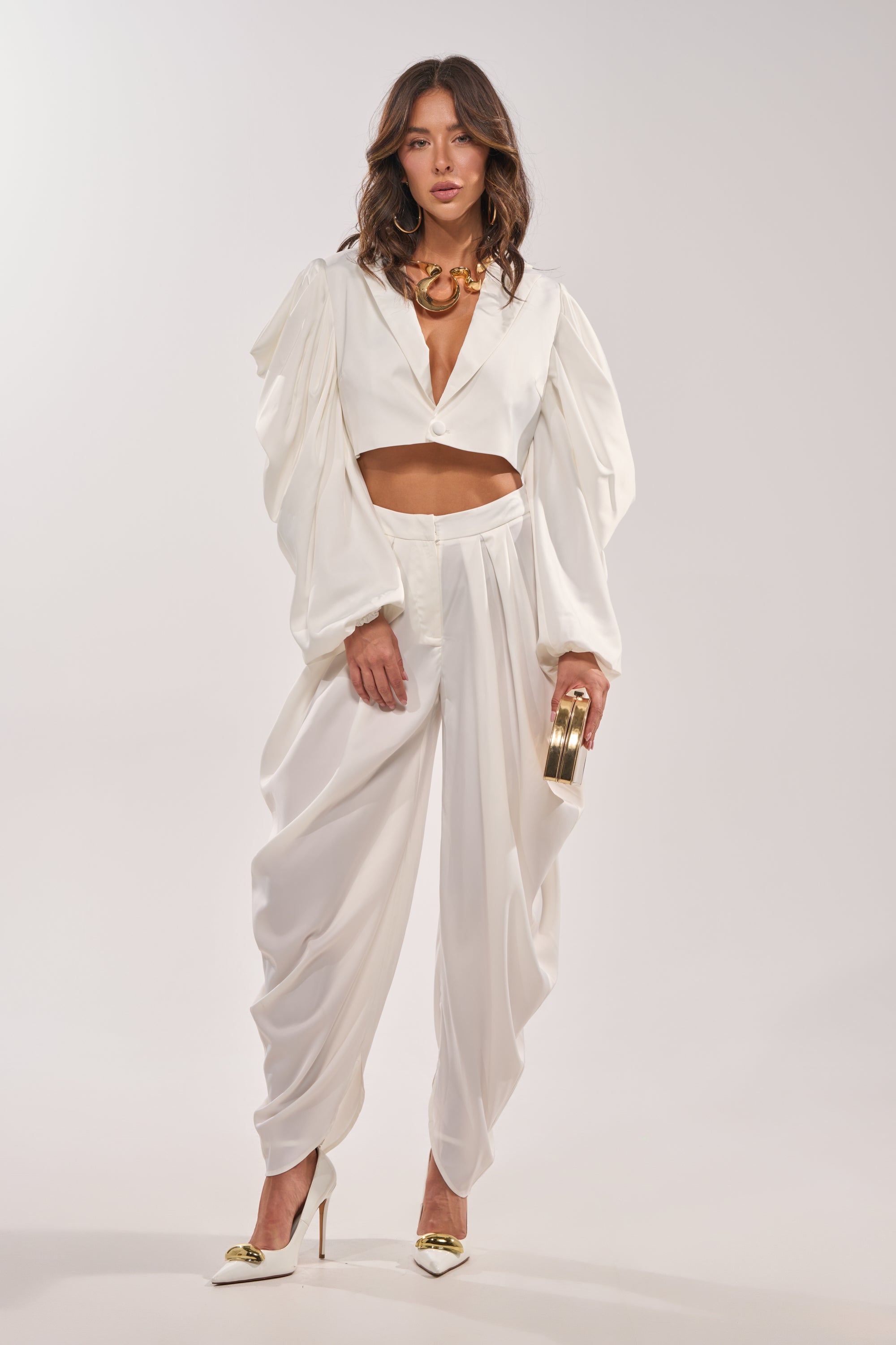 A woman models the NEW BEGINNING CROPPED BLAZER, a chic white style with cape sleeves, paired with wide-leg pants. She accessorizes with a gold clutch, gold heels, and a chunky gold necklace against a plain background. 6