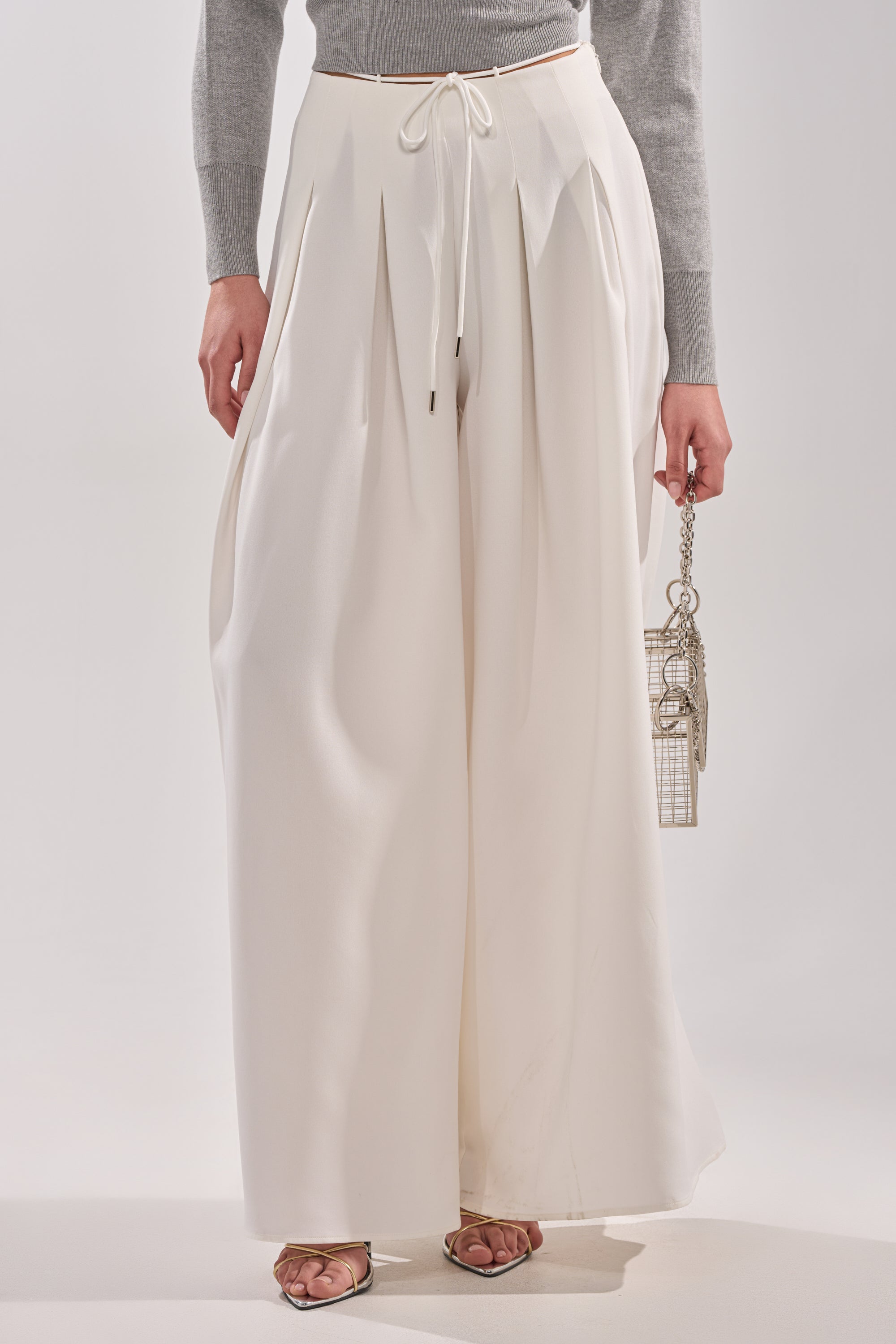 Wearing the GLORIOUS PLEATED WIDE LEG TROUSER—high-waisted, white with a drawstring waist—paired with a gray long-sleeve top, heeled sandals, and a metallic handbag creates an effortlessly chic, versatile look.
