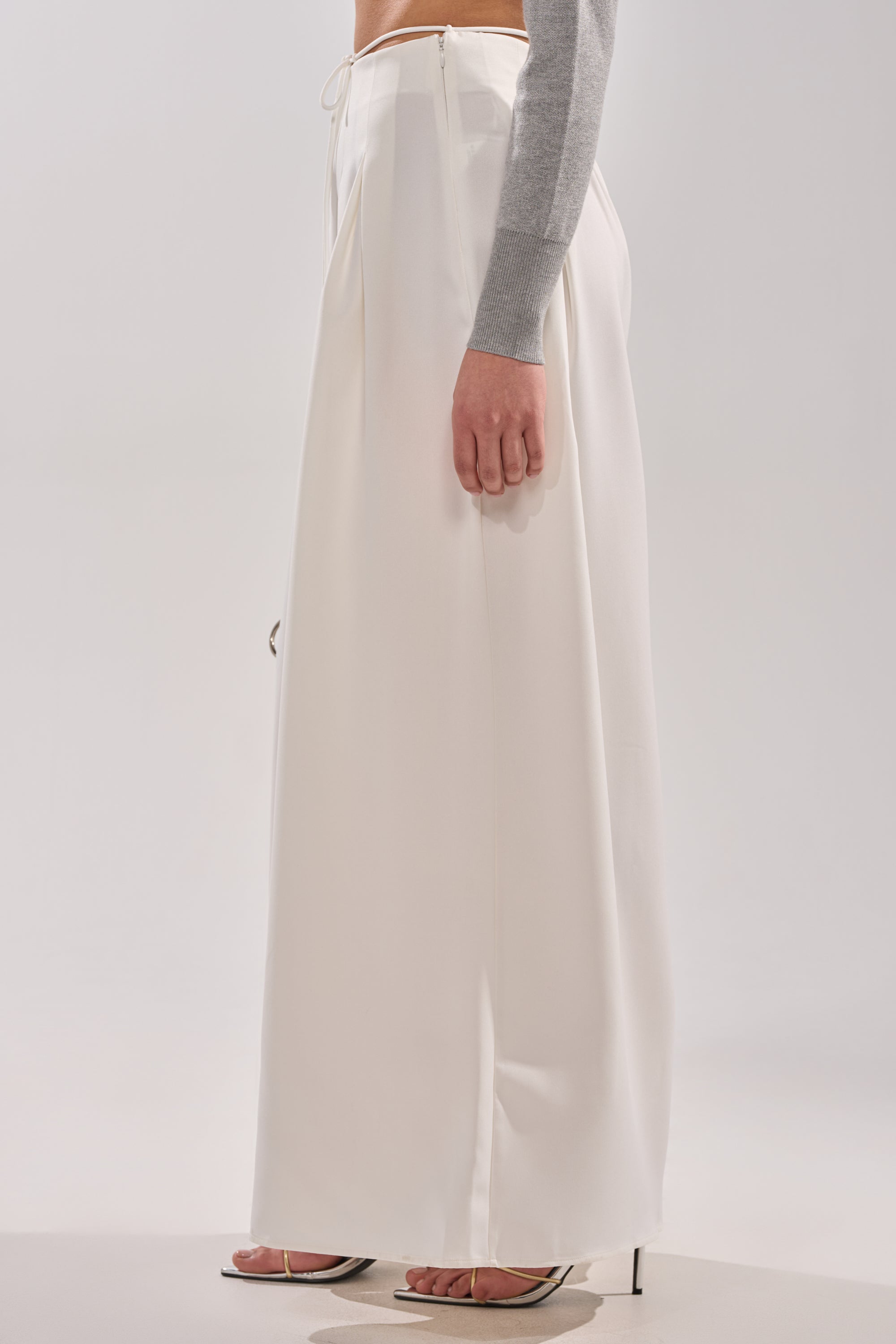 Dressed in the GLORIOUS PLEATED WIDE LEG TROUSER—high-waisted and white—with a light gray long-sleeve top and open-toe heeled sandals, a person stands sideways against a plain light background. Photo is mid-torso down.