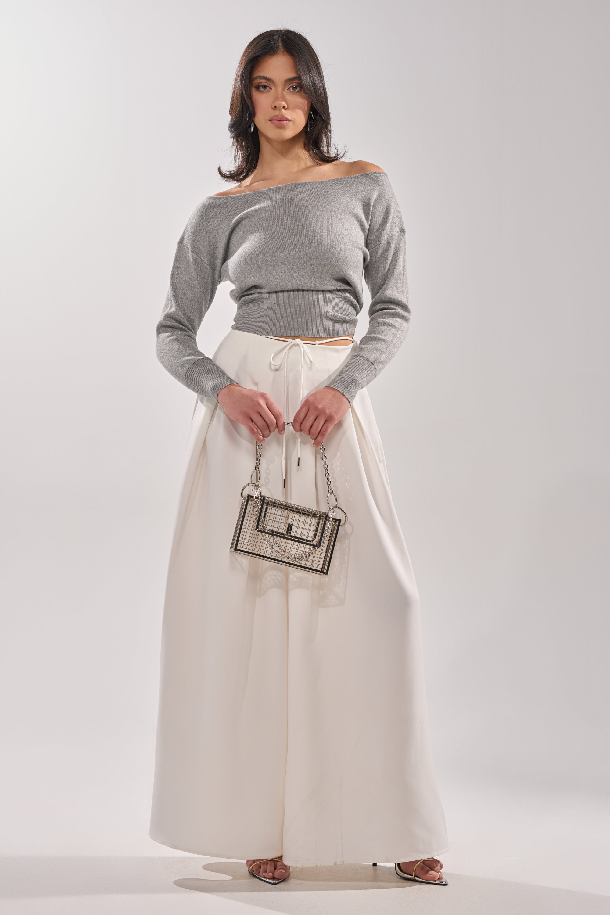 A woman poses against a light background in a gray off-the-shoulder sweater and the GLORIOUS PLEATED WIDE LEG TROUSER—essential white wide-leg pants. She holds a clear handbag and looks confidently at the camera in heeled sandals.