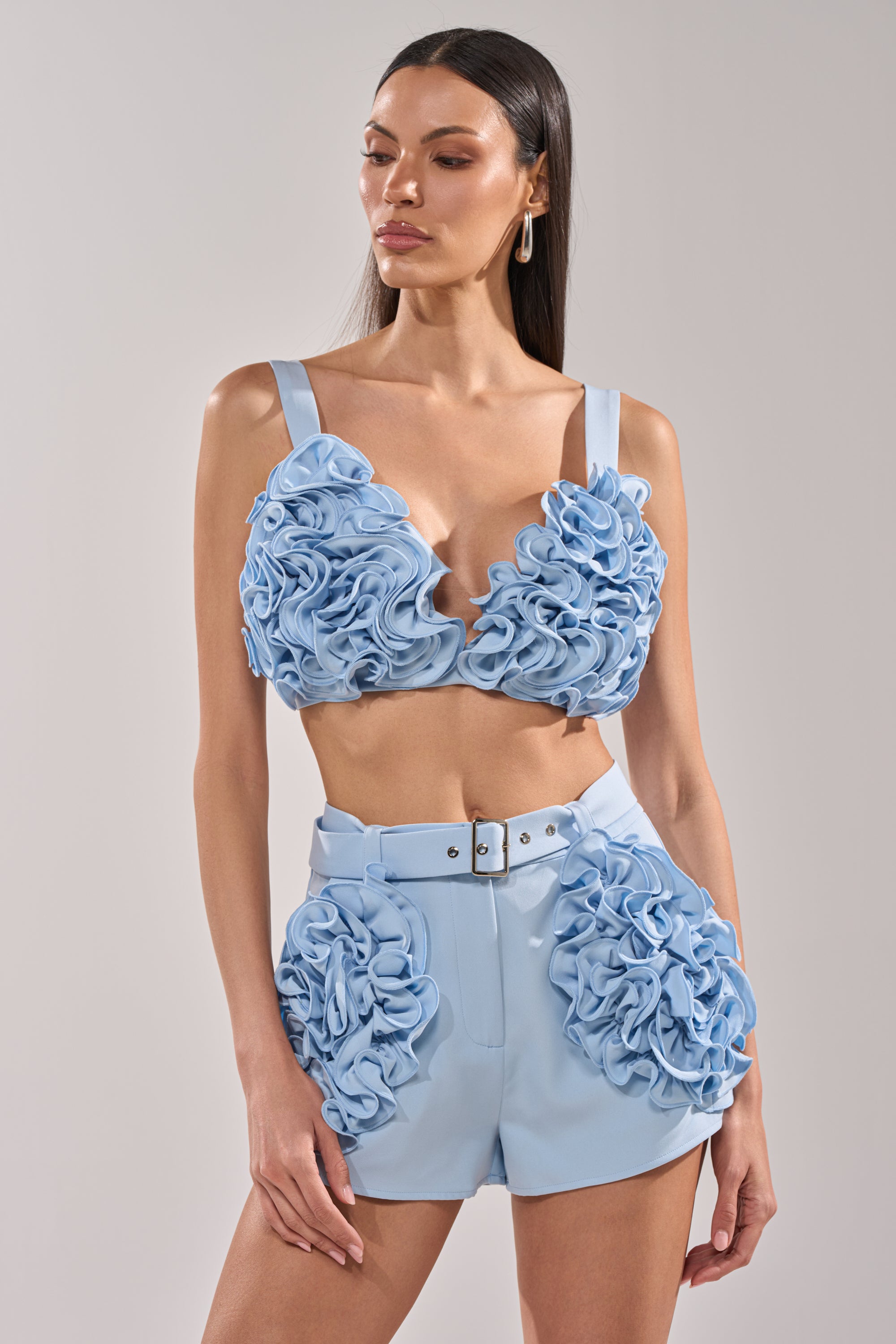 A woman wears the FLOWER GIRL BRALETTE, a baby blue coordinated set featuring 3D floral appliqués on the bralette and high-waisted shorts, standing against a plain background. 0