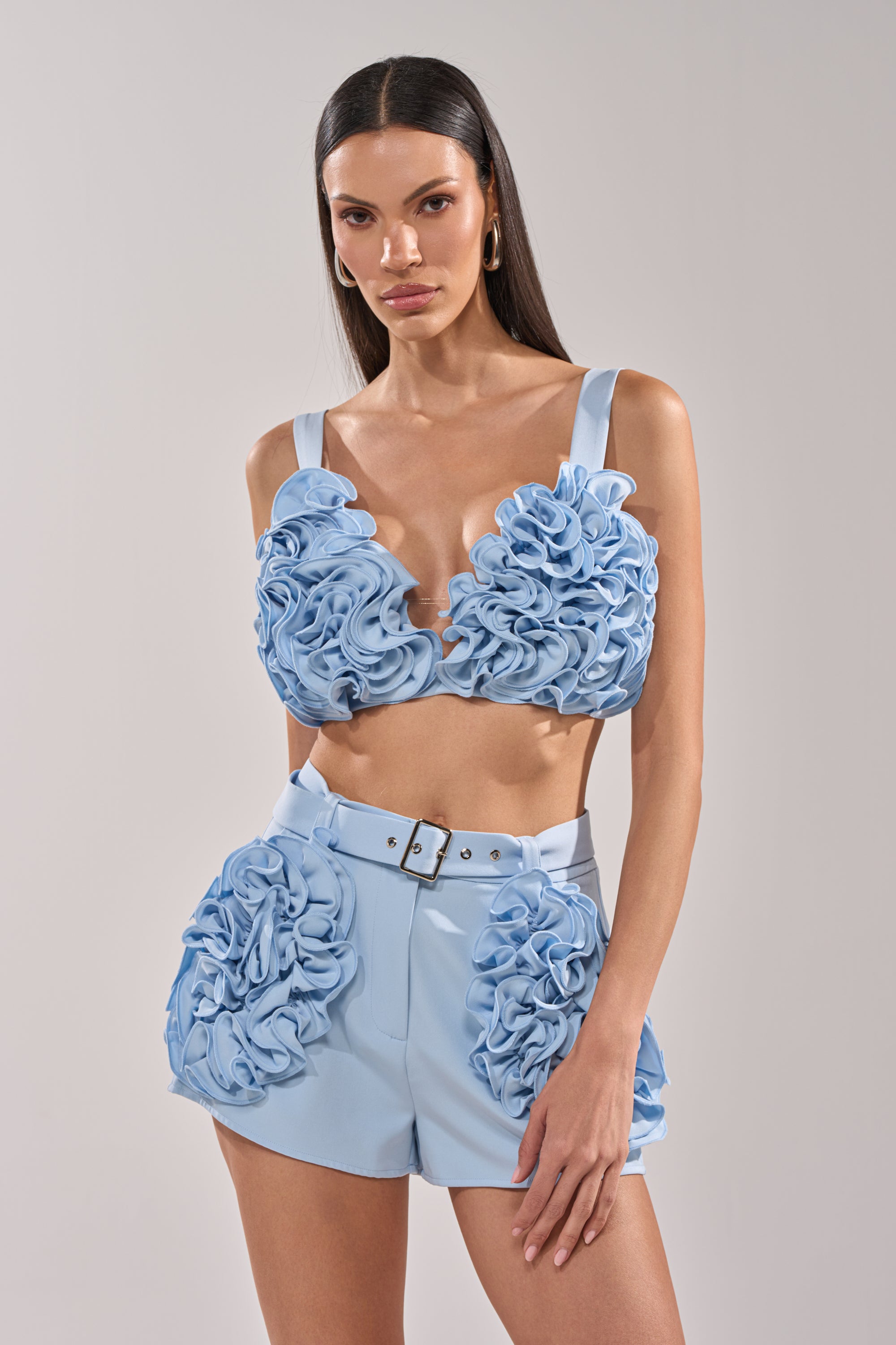 A woman faces the camera wearing the FLOWER GIRL BRALETTE in baby blue with matching high-waisted shorts, both featuring large 3D floral appliqués and a cropped fit. She has long, straight hair and a neutral expression. 1