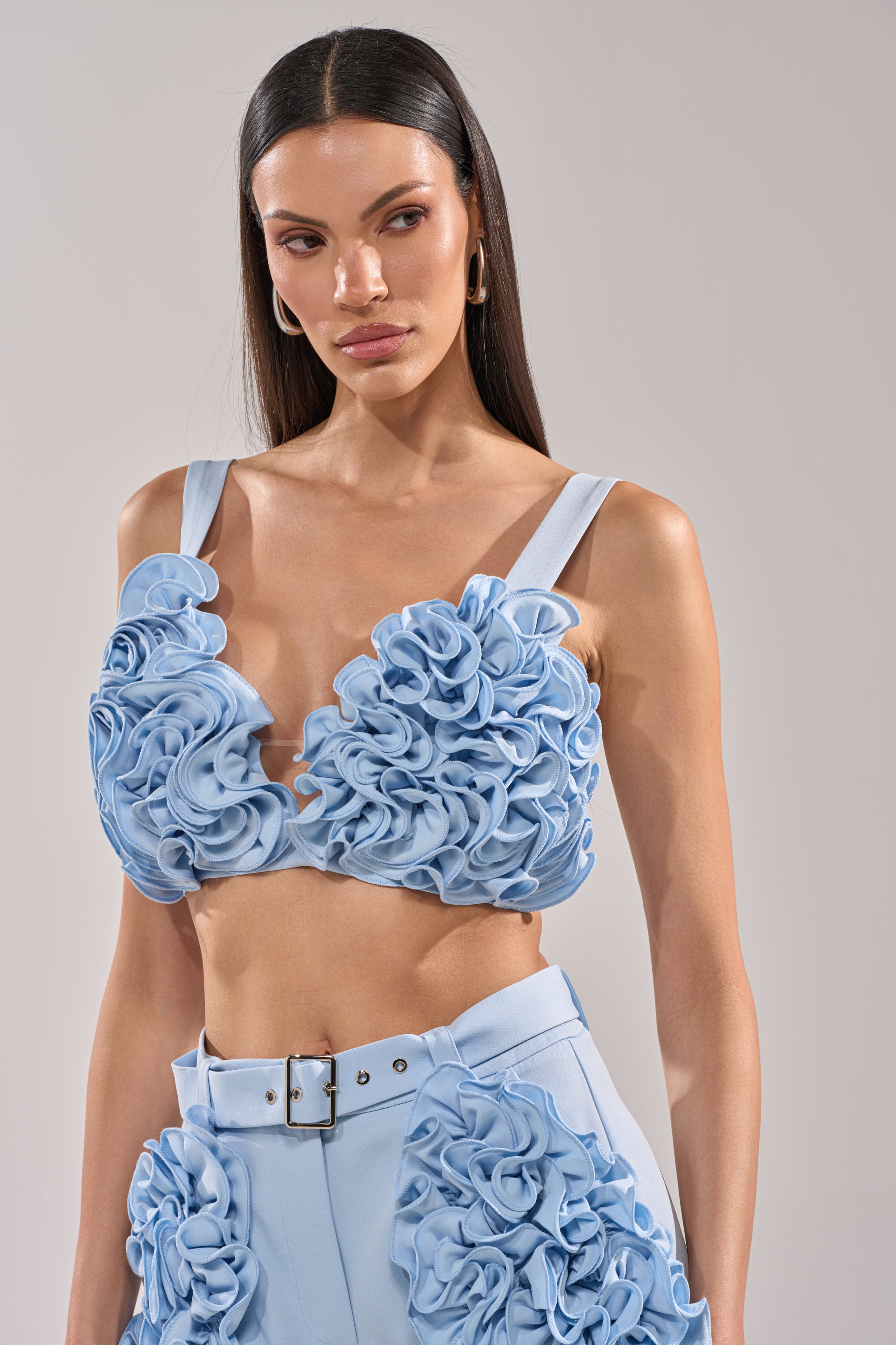 A woman with long, straight dark hair wears the FLOWER GIRL BRALETTE, a baby blue bralette with three-dimensional ruffle detailing and a cropped fit. She pairs it with high-waisted pants and poses against a plain background. 2