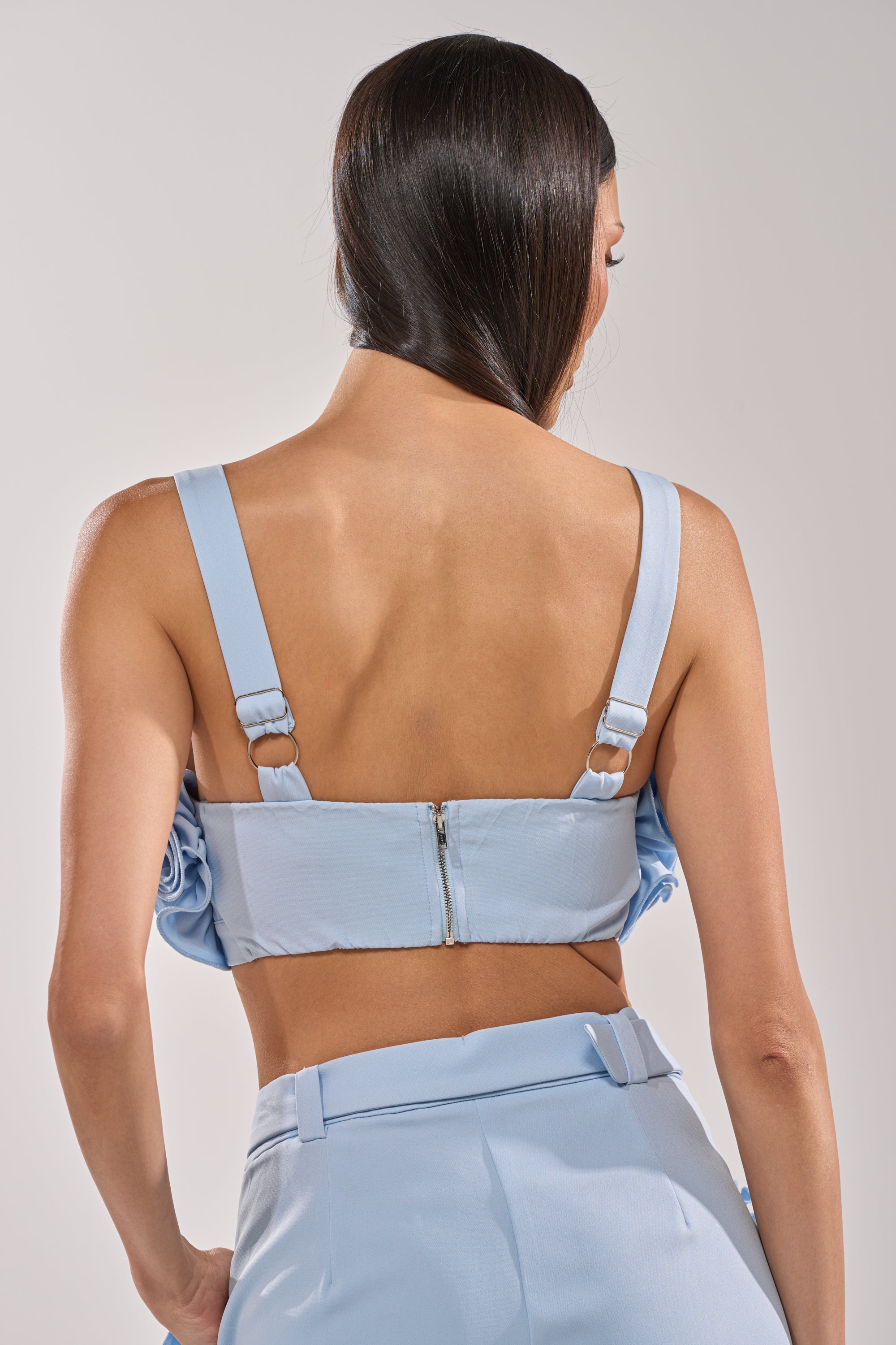 A woman with long, straight brown hair wears the supportive FLOWER GIRL BRALETTE in baby blue and high-waisted pants, photographed from the back against a plain light background. 4