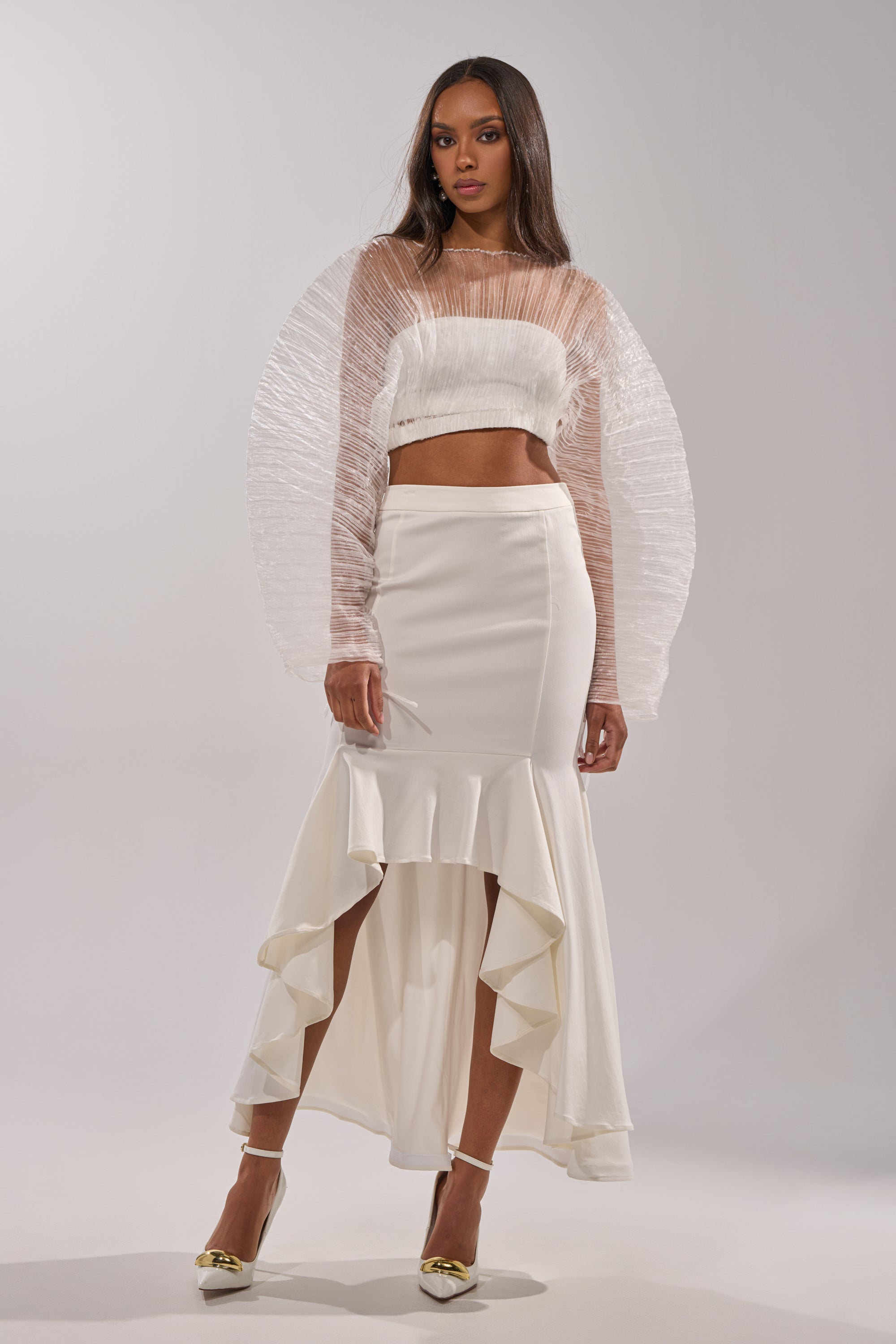 A woman wears the LEMON SATIN MAXI SKIRT with a sheer, textured white long-sleeve crop top and pointed-toe heels, posing against a plain light background. 0