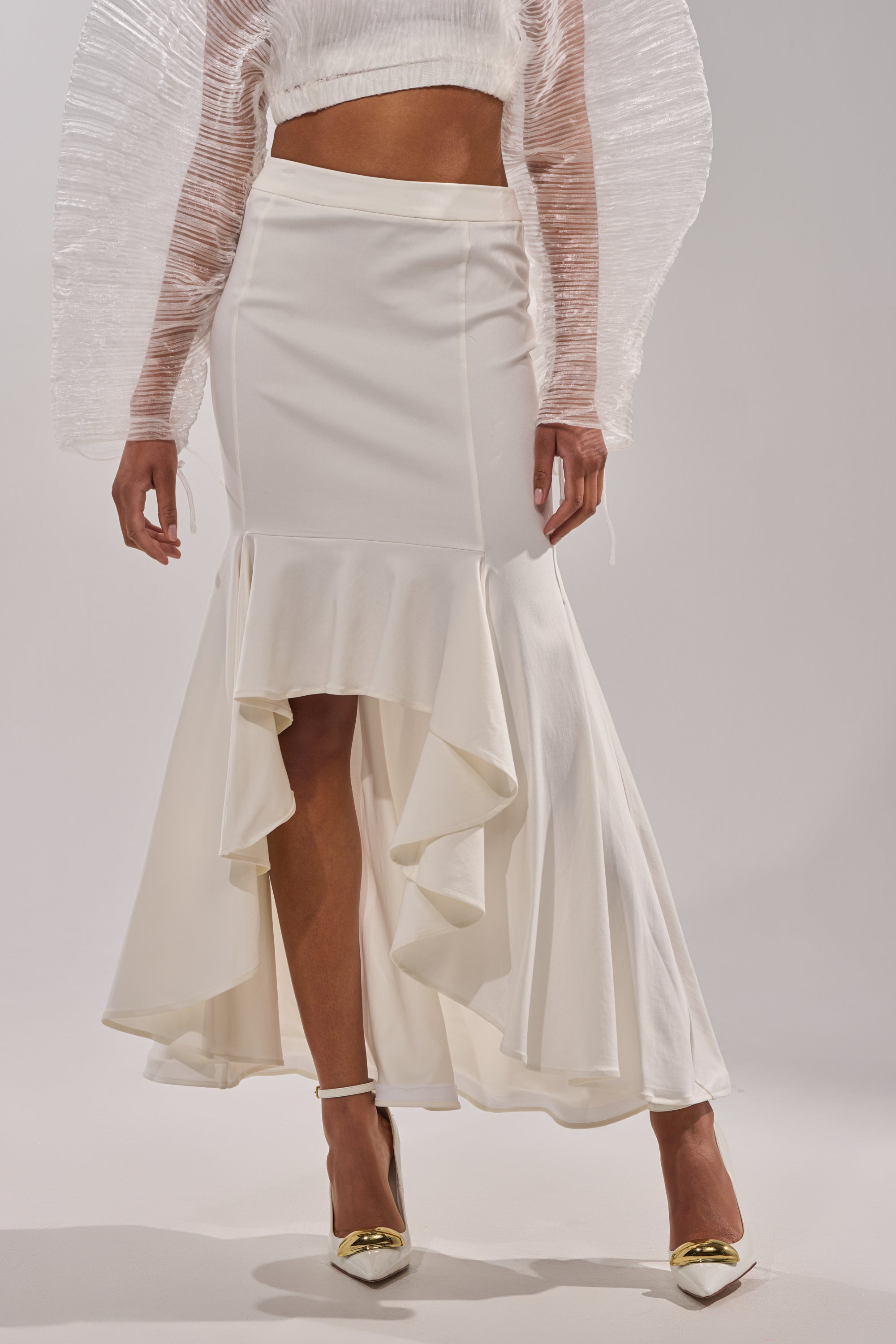 A person wears the LEMON SATIN MAXI SKIRT, a high-waisted, white mermaid skirt with a ruffled high-low hem, styled with a textured long-sleeve white top and white heels featuring gold accents. The image is cropped at the chest. 1