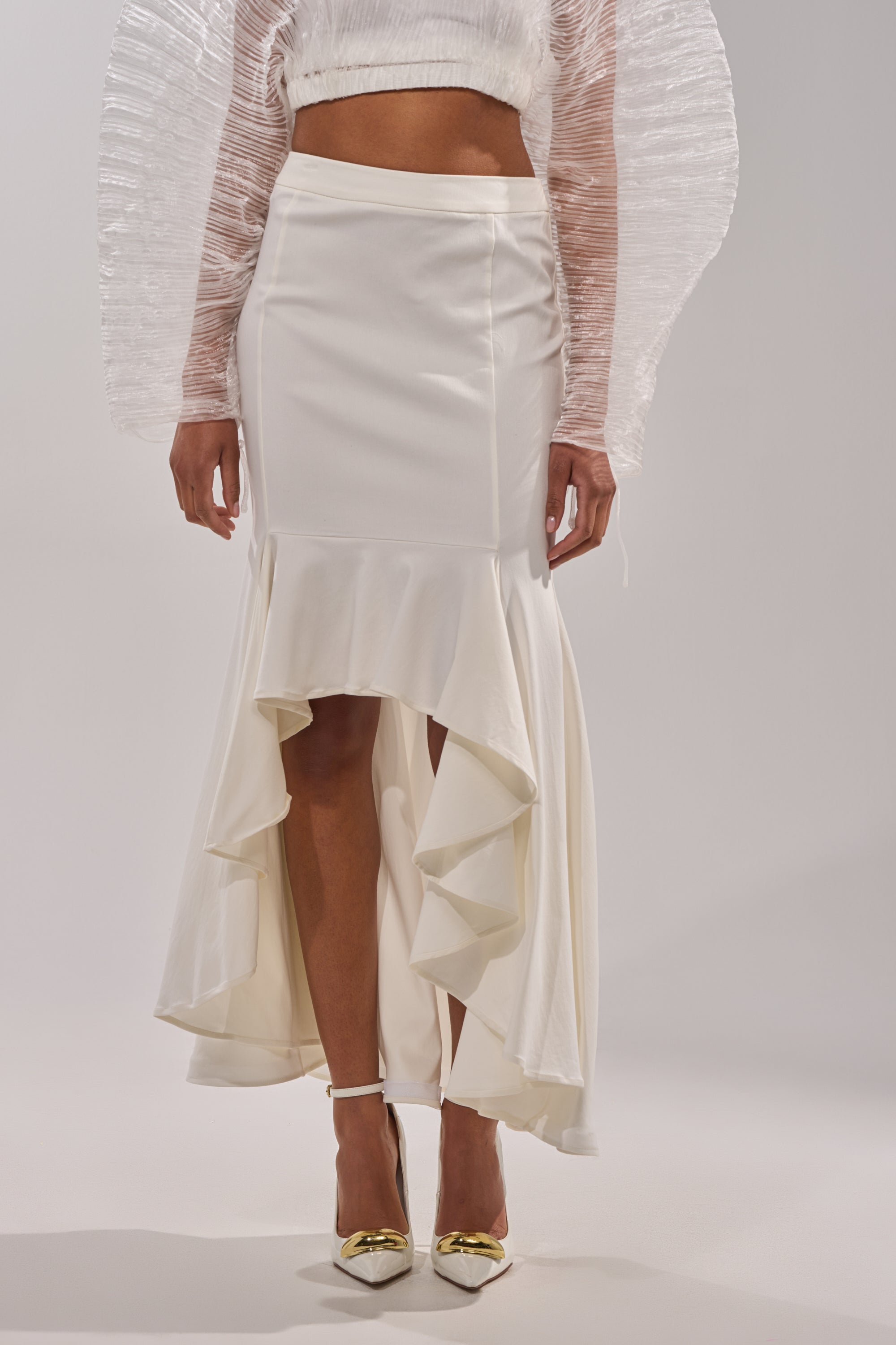 A person stands against a light gray background wearing the LEMON SATIN MAXI SKIRT, styled with layered ruffles, a fitted waist, sheer long-sleeved top, and white pointed heels with gold accents. 2