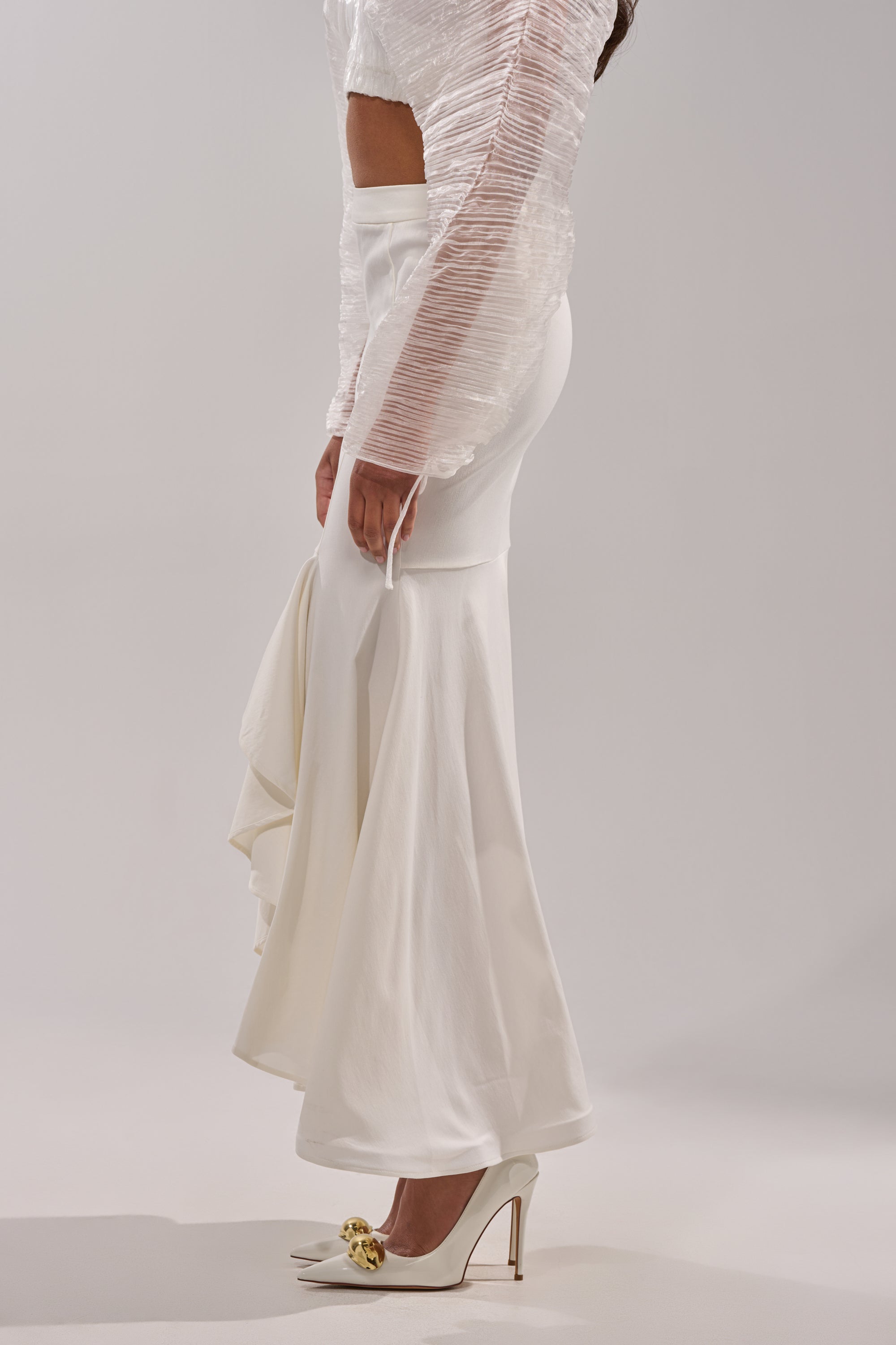A woman stands against a plain light background wearing a textured, long-sleeve crop top and the LEMON SATIN MAXI SKIRT, paired with pointed white heels featuring gold embellishments. 3