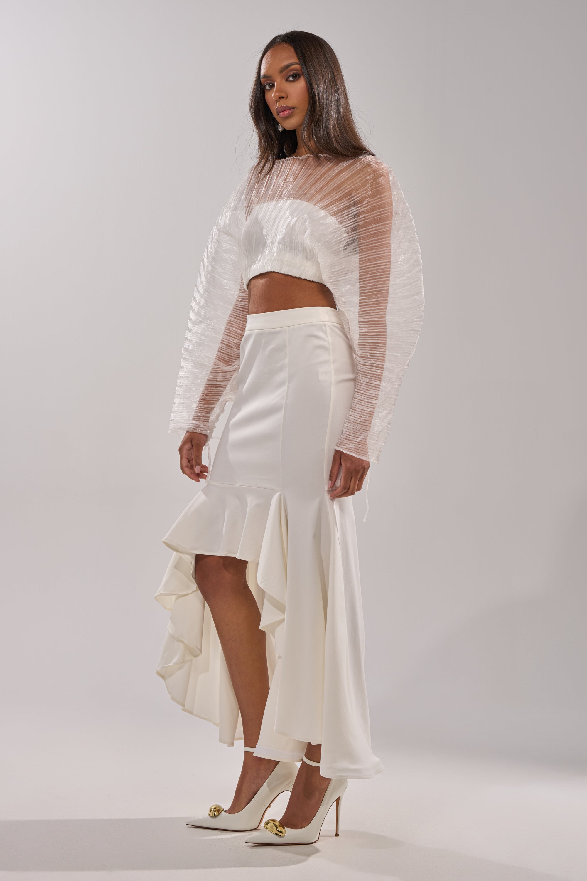 A woman poses against a plain background wearing the LEMON SATIN MAXI SKIRT featuring asymmetrical ruffled details, styled with a sheer long-sleeve cropped top and white high heels for an elegant look. 5