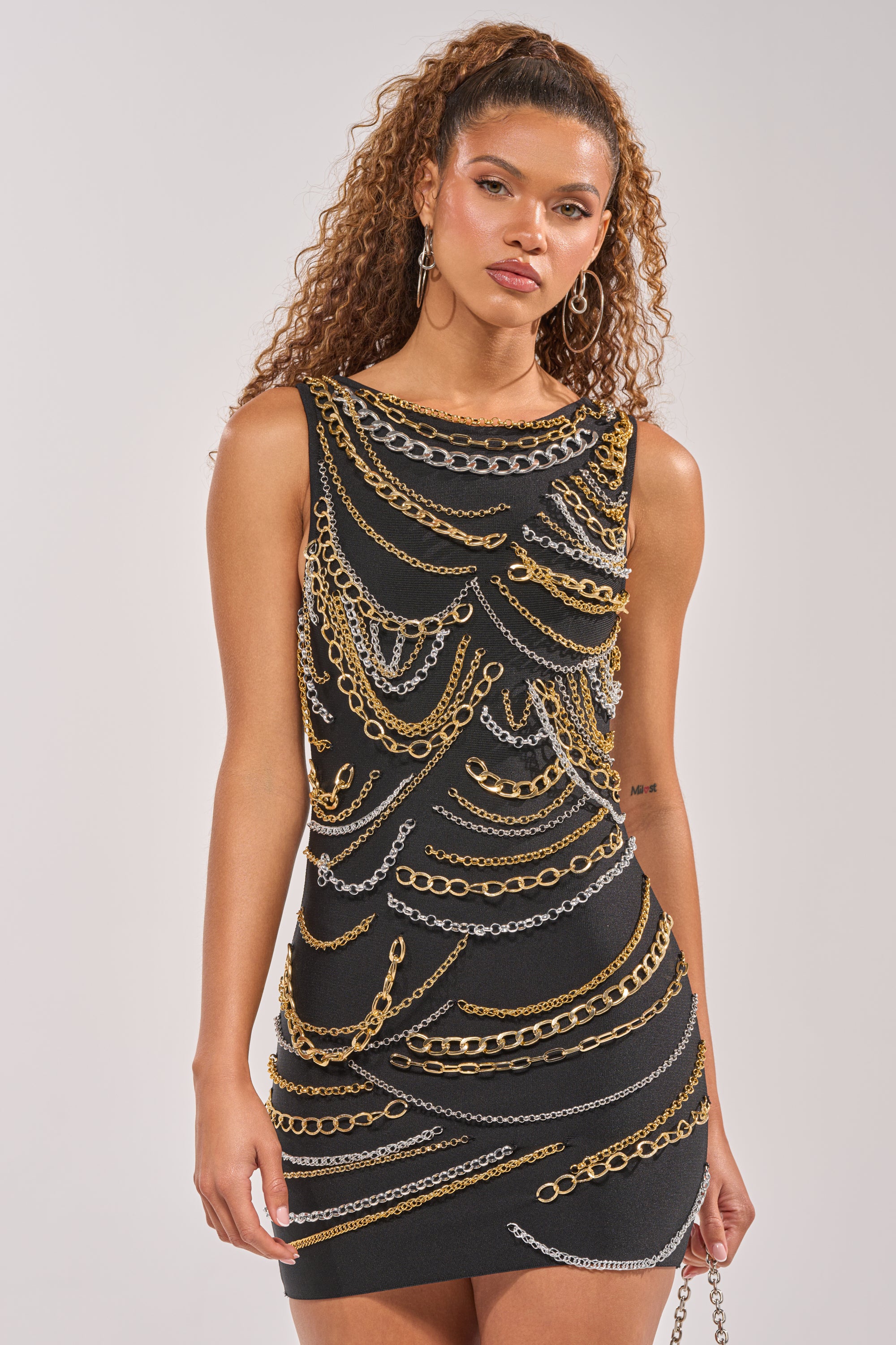 A woman wears the BLOWING KISSES CHAIN DETAIL MINI DRESS with layered gold and silver chains. Standing against a light backdrop, she sports curly hair and hoop earrings—a confident, chic look for festivals. 1
