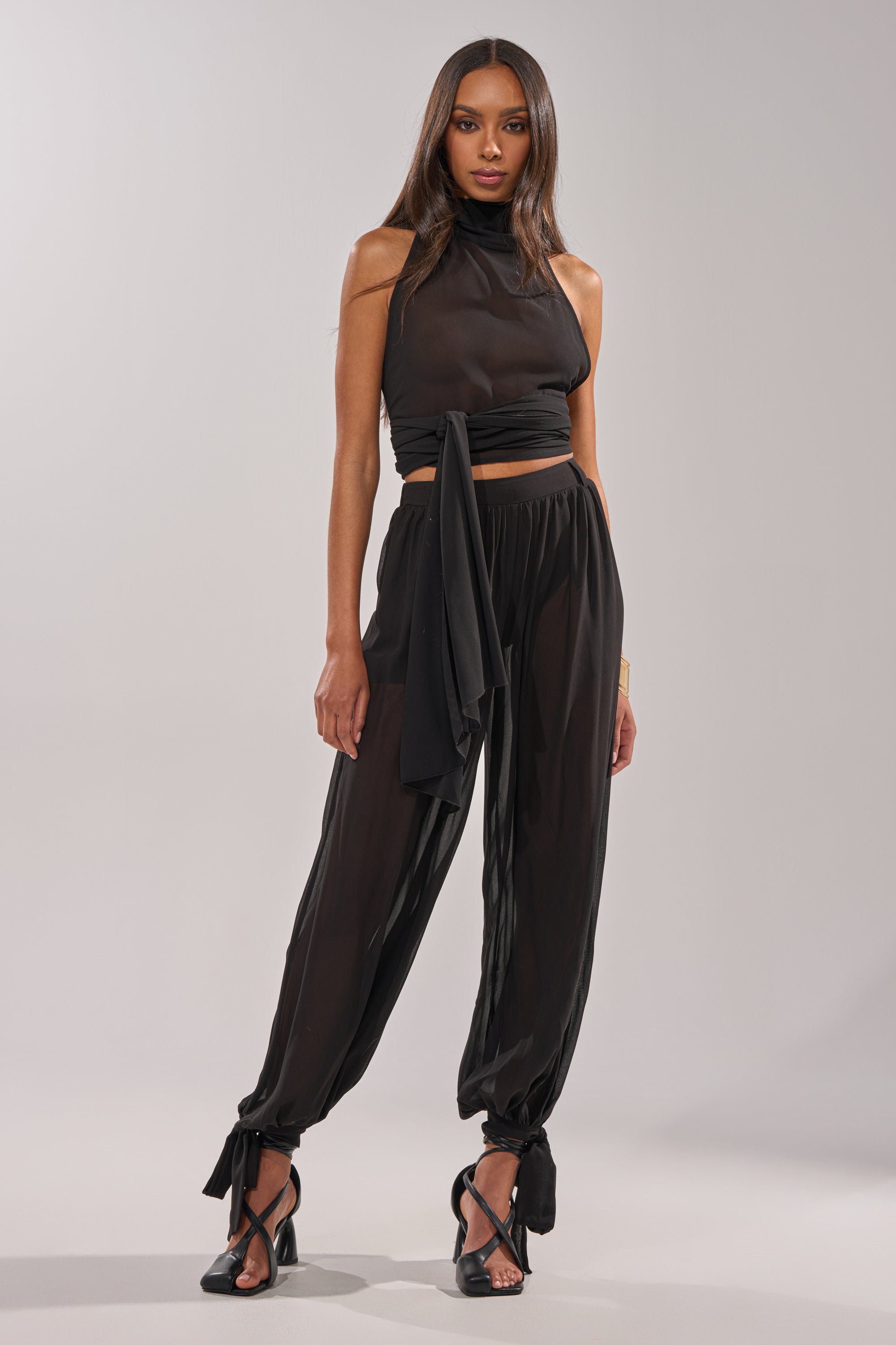 A woman poses confidently against a plain light background, wearing the TRENDY BALLON PANT tied at the ankles with a sheer black sleeveless high-neck top and black heeled sandals. 3