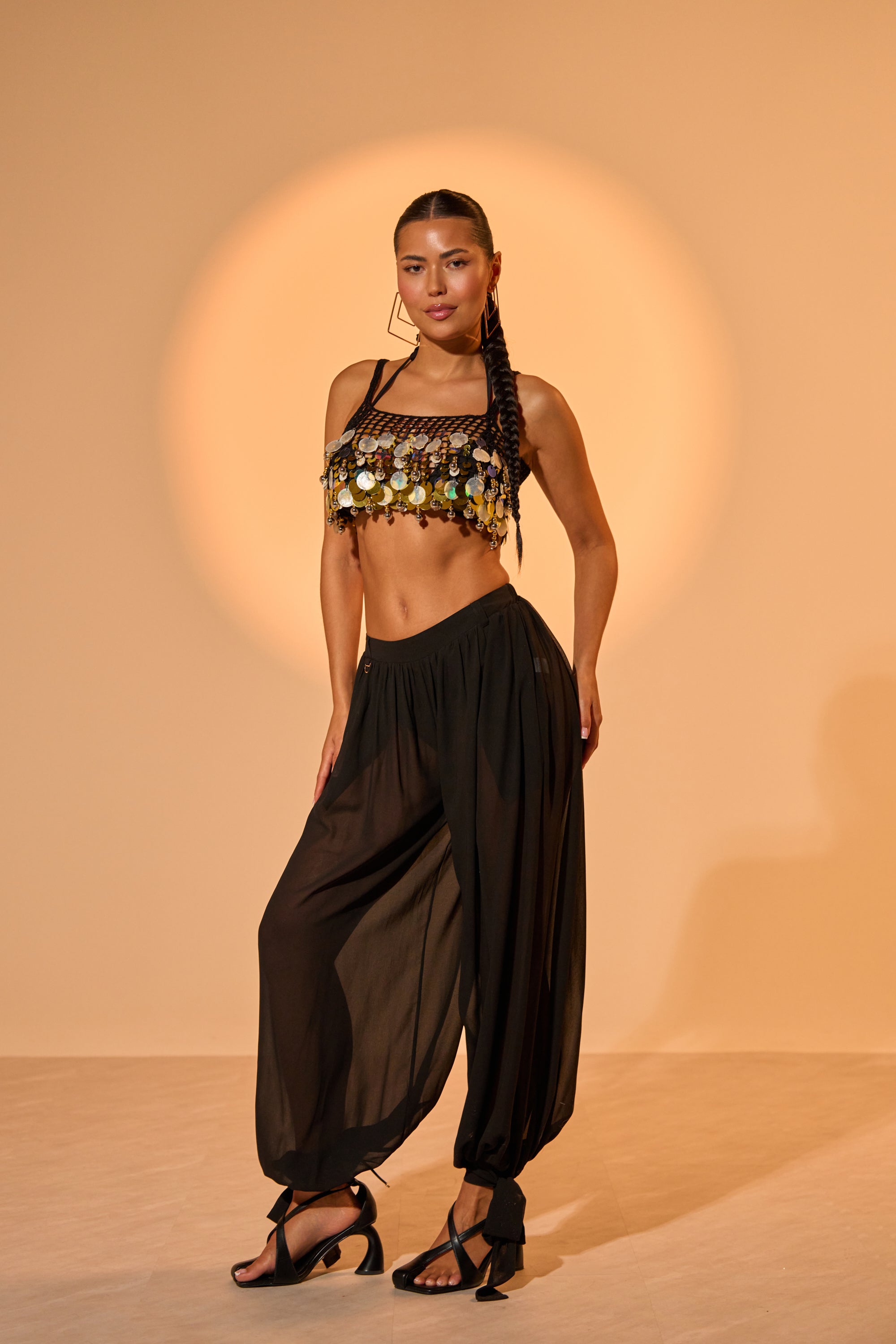 A woman stands confidently in front of a warm beige backdrop, wearing the TRENDY BALLON PANT in sheer black chiffon with a gold-coin crop top, sandals, and long braided hair. 0