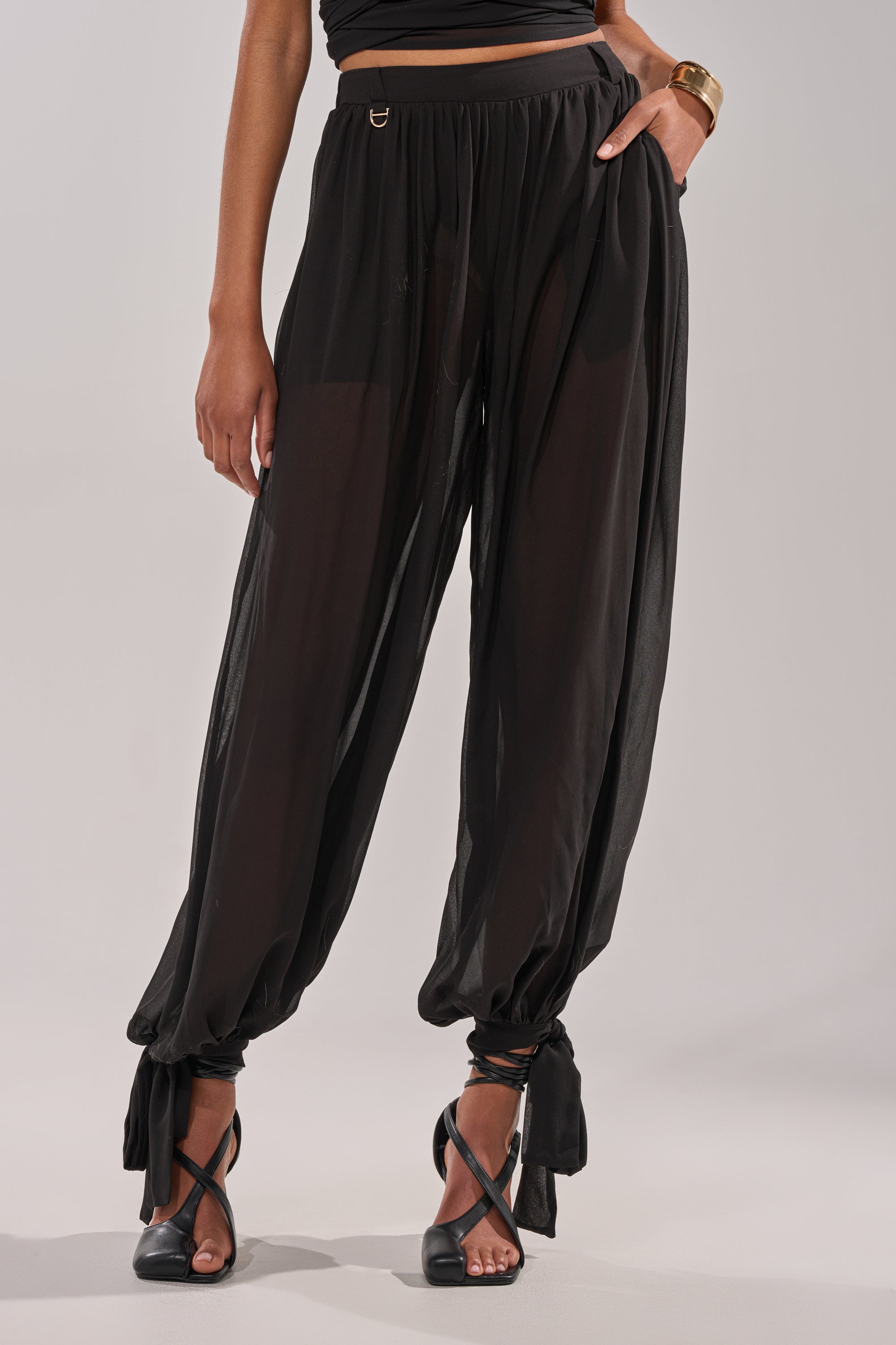 A person stands against a plain light background wearing the TRENDY BALLON PANT—high-rise chiffon pants with ankle tie details—paired with black high-heeled sandals, a gold bracelet, and a black crop top. 4