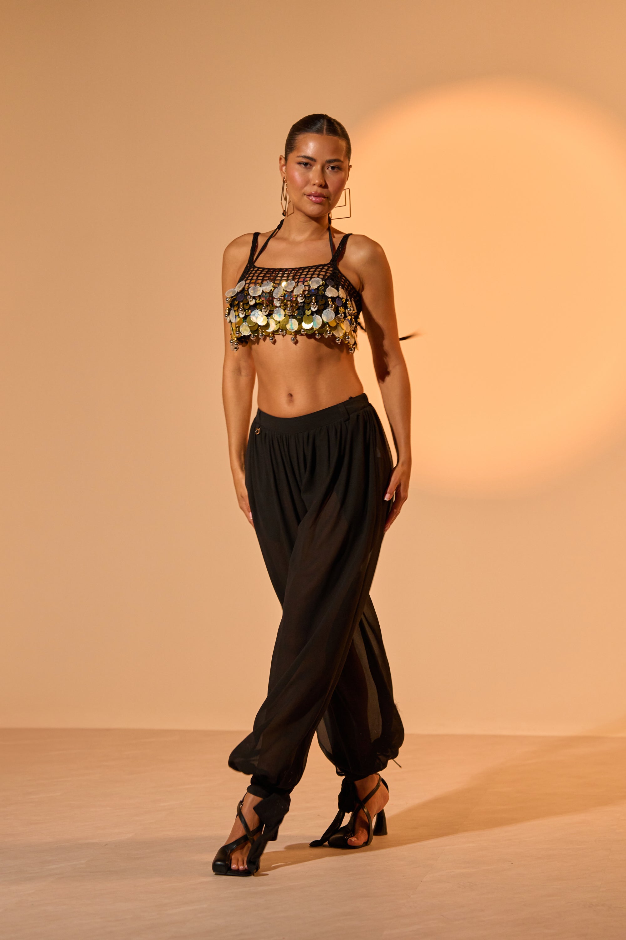 A woman poses confidently in a gold coin-embellished crop top, TRENDY BALLON PANT styled as black high-rise chiffon balloon pants, and black heels against a beige background with a circular light pattern. 1