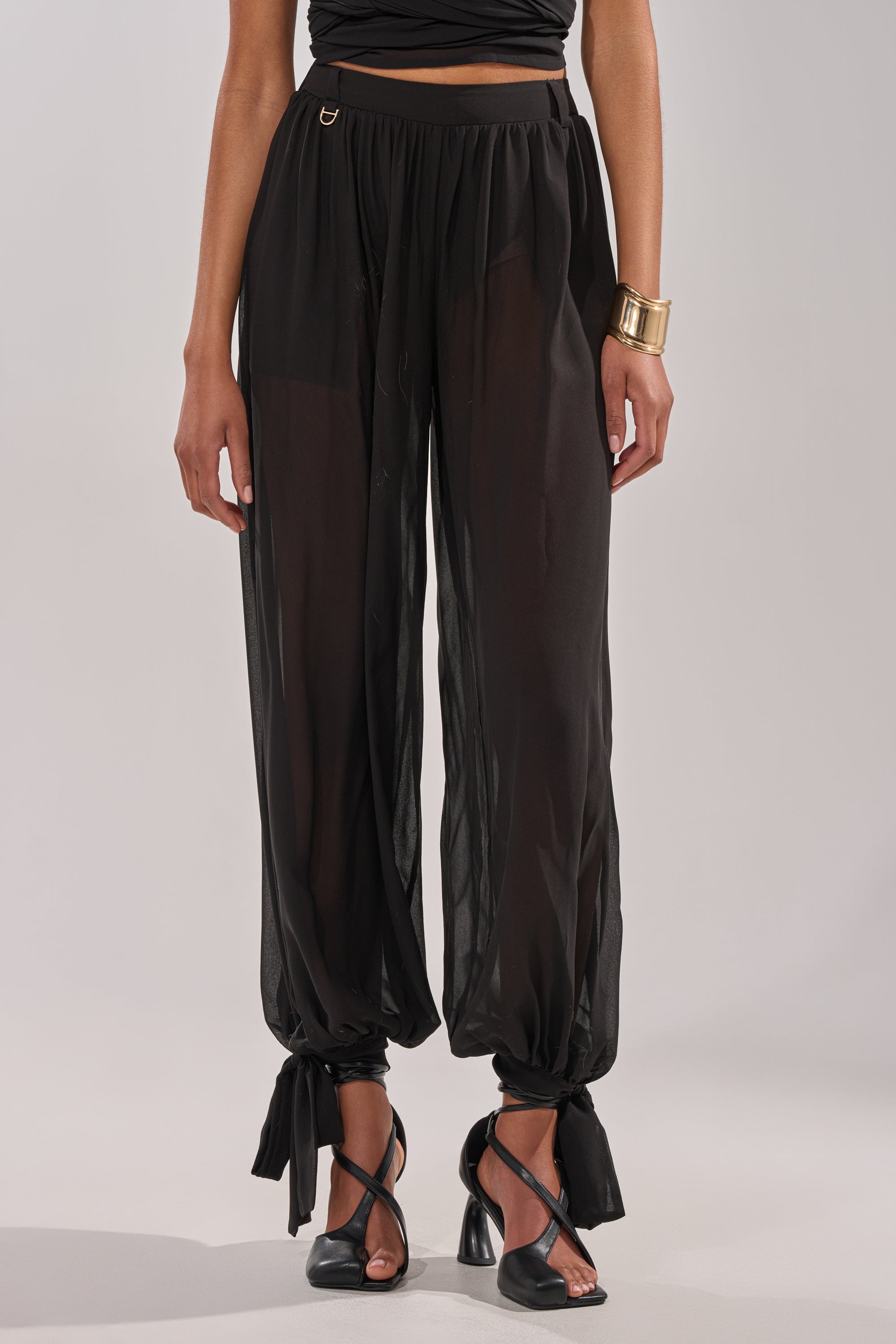 A person wearing TRENDY BALLON PANT in sheer black chiffon with ankle ties, paired with black heeled sandals, a black top, and a large gold cuff bracelet, stands against a neutral background. 5